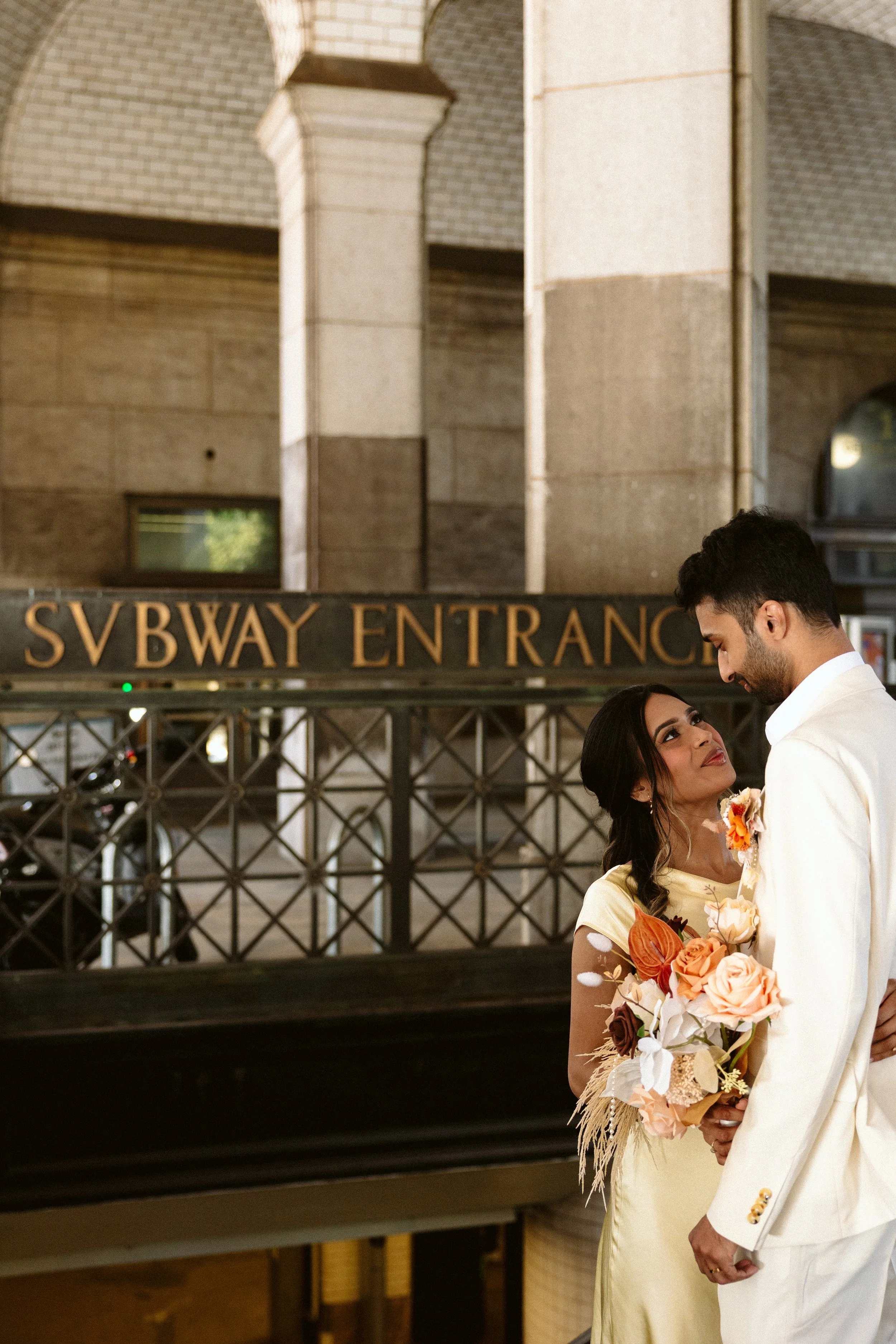 A couple in wedding attire, looking at each other and holding a bouquet of flowers, standing near a subway entrance with an arch and a sign that reads 'SUBWAY ENTRANCE'.