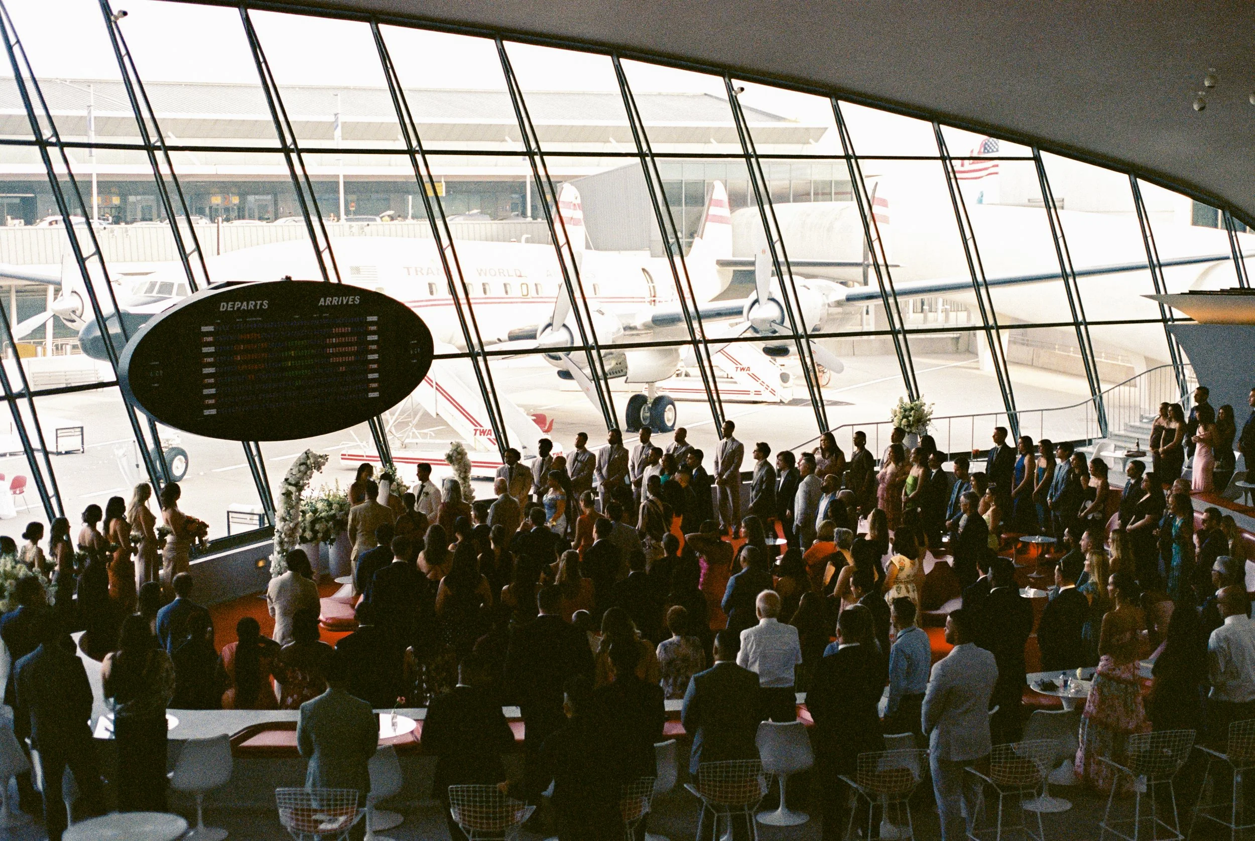 Indoor view of a wedding ceremony at the TWA hotel in JFK airport with large glass windows showing airplanes outside. Inside, a crowd of people stands and sits around tables observing.