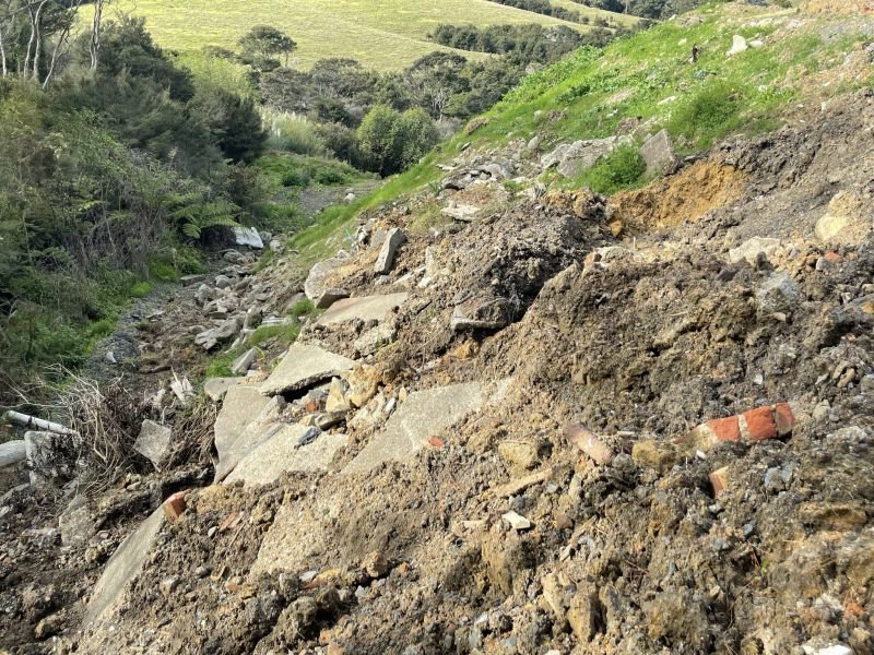 SQN GeoSciences Disposal to Land Risk Management Slope with broken concrete and dirt, signs of a landslide or erosion, with green grassy hills and trees in the background.