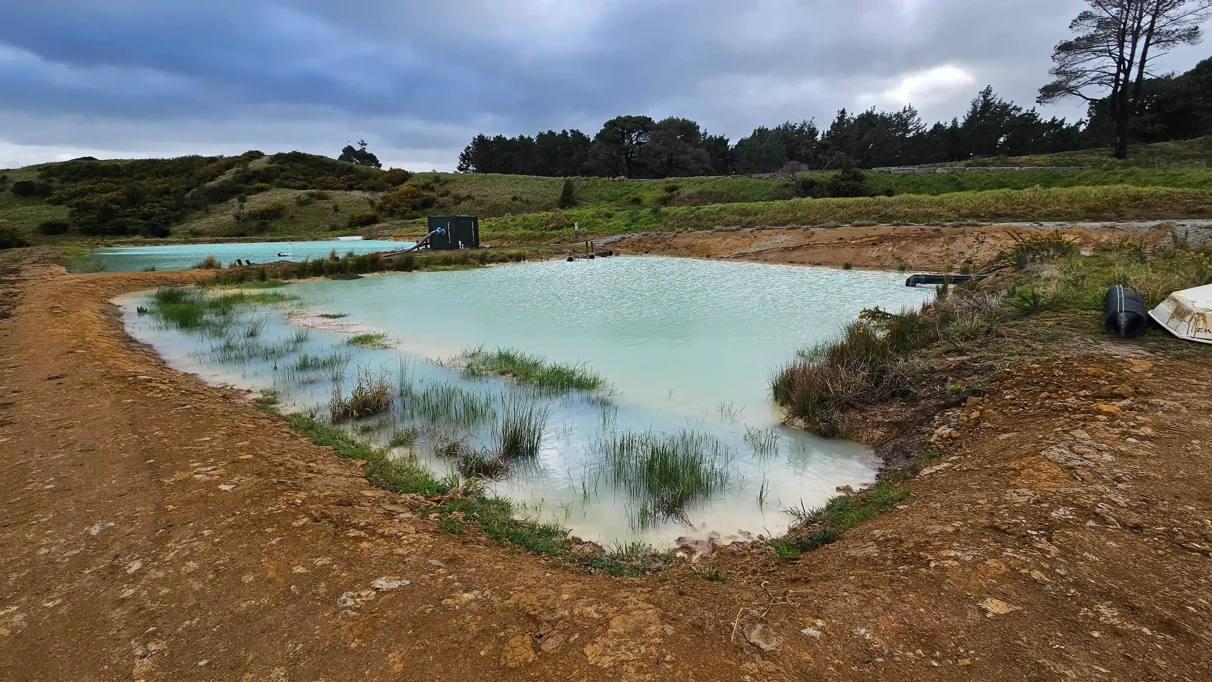 SQN GeoSciences Image A geothermal hot spring pool with milky blue water surrounded by brown dirt and grass, with a cloudy sky overhead and hills of trees and shrubs in the background.