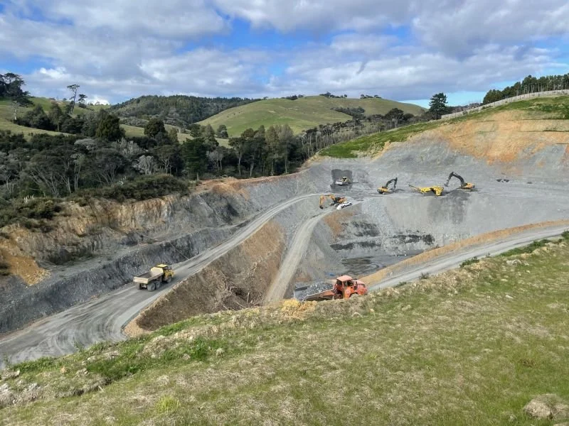 View of a large construction site in a hilly area with heavy machinery, including excavation equipment, working on a large open pit. Green hills and trees are in the background under a partly cloudy sky.