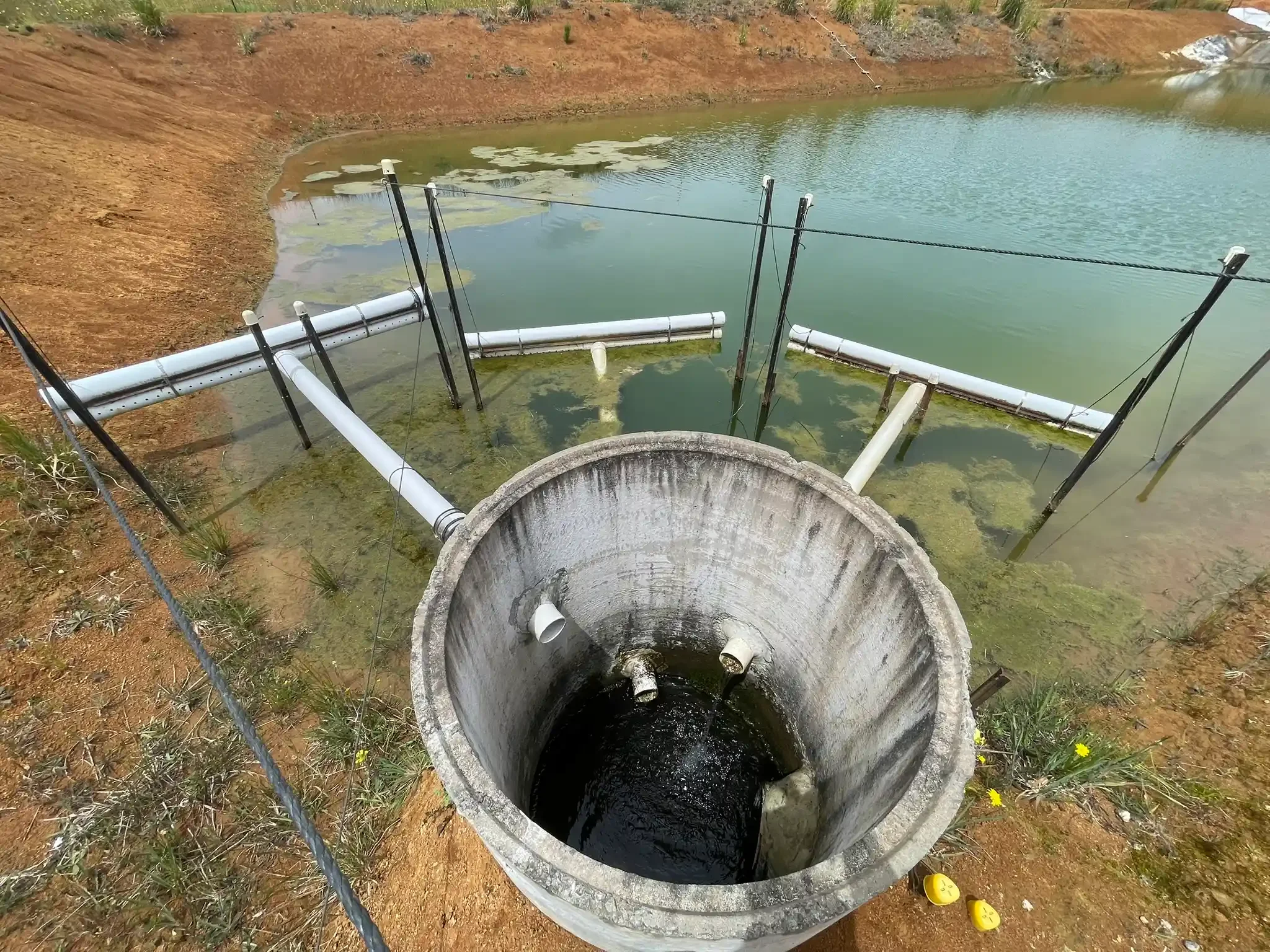 SQN GeoSciences Compliance Monitoring A water reservoir with a concrete outlet pipe funneling water into a pond, surrounded by a dirt embankment and algae-covered water.
