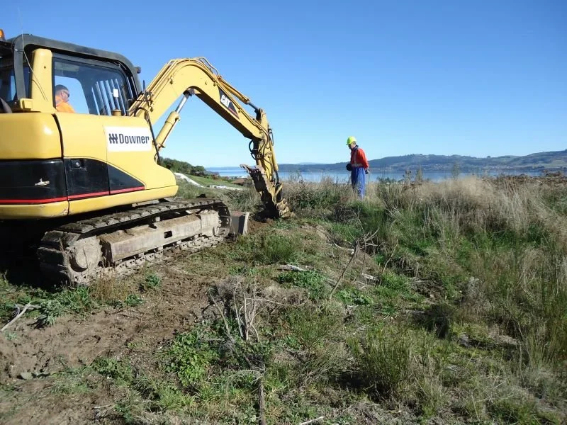 A yellow excavator working on a grassy shoreline, with a construction worker in safety gear nearby, and a body of water with hills in the distance under a clear blue sky.
