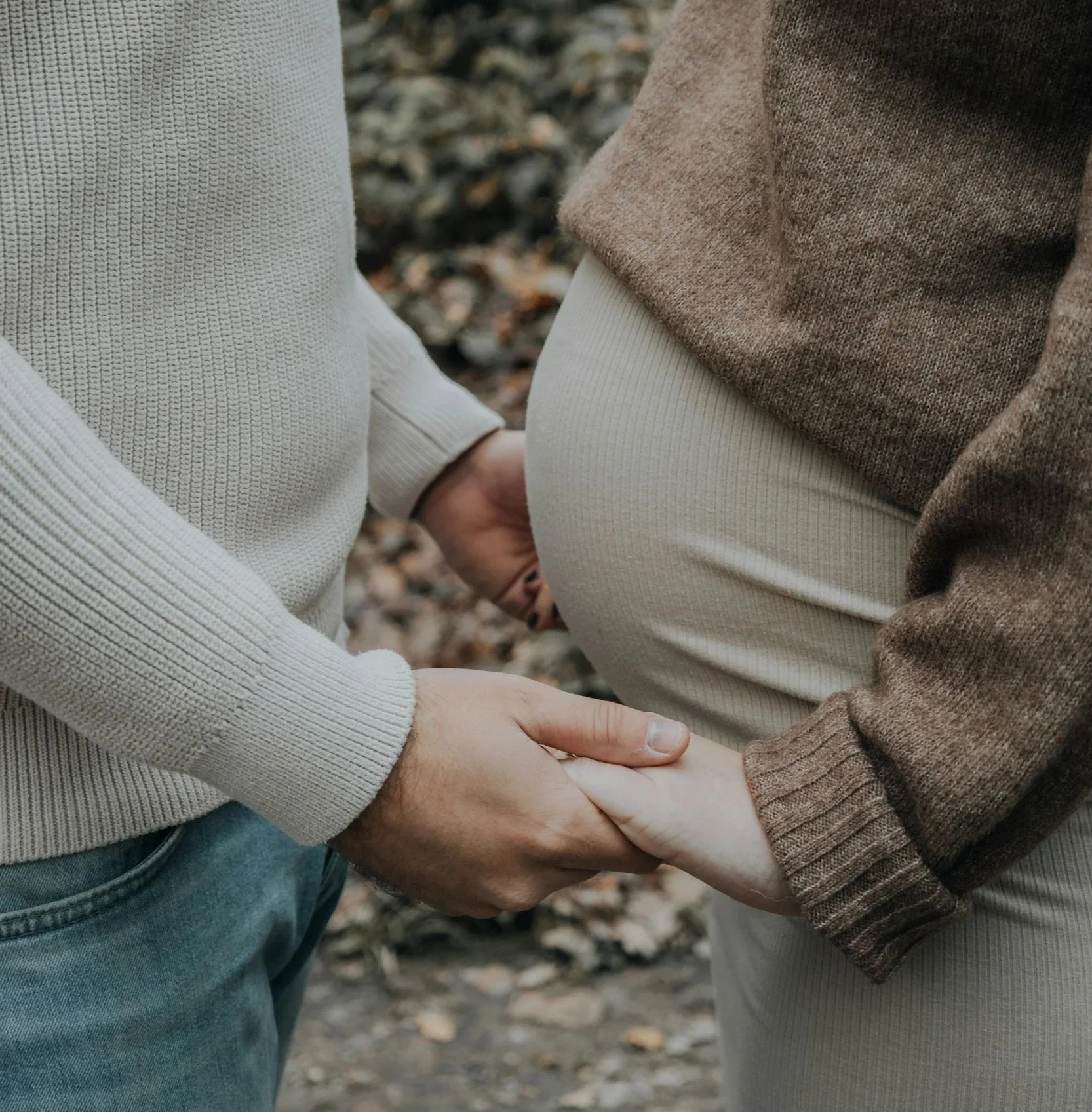 A close-up of a couple holding hands, with the focus on their hands and forearms, standing outdoors with fallen leaves in the background.