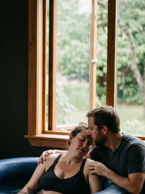 A man and a woman embrace in a small indoor pool near a window showing green trees outside, with the woman resting her head against the man's forehead.