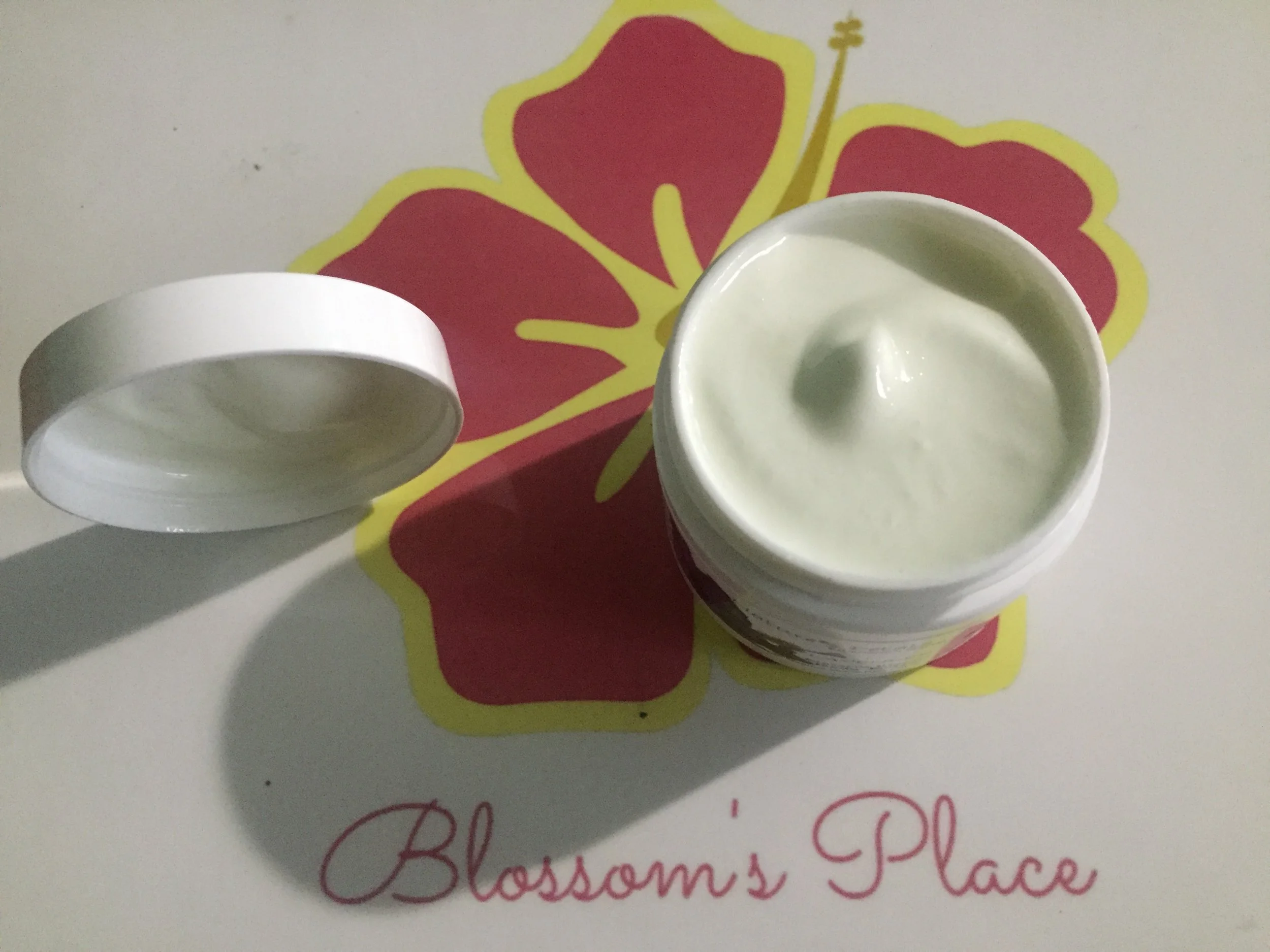 Open container of white cream or lotion on a surface with a floral design and the words 'Blossom's Place' written underneath.