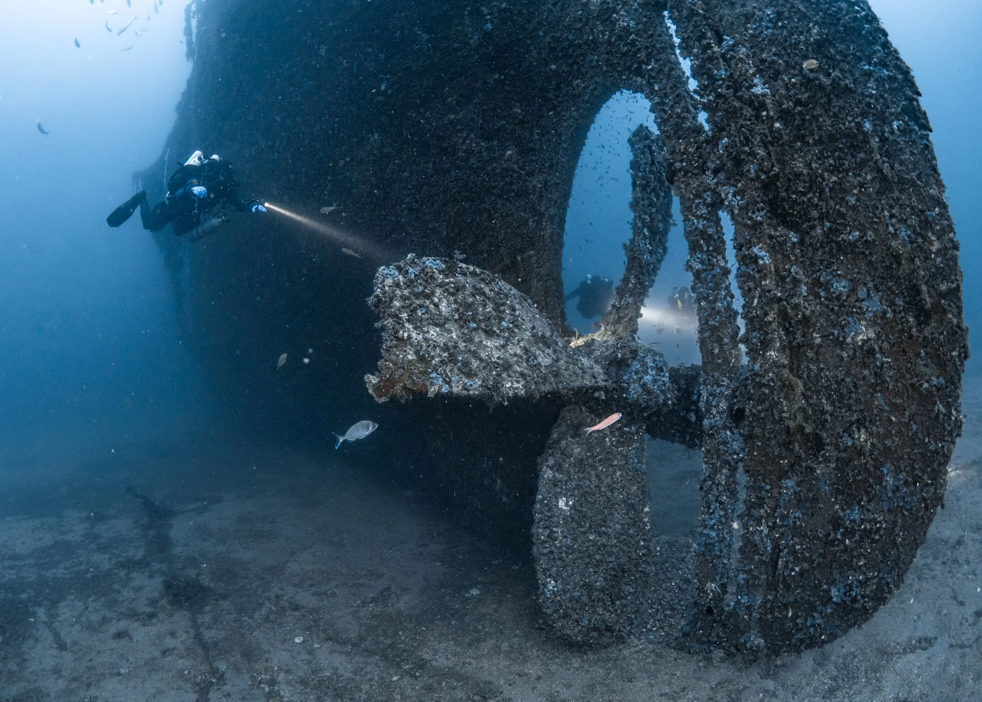 Technical divers exploring a massive shipwreck propeller covered in marine growth in deep blue water of south Sardinia.