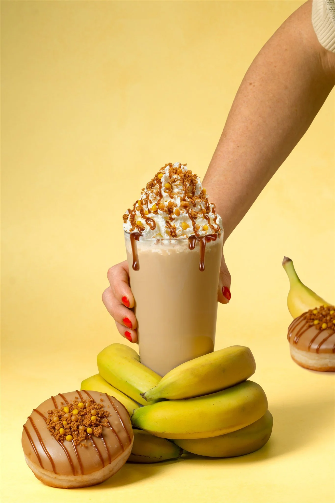 A hand with red nail polish holding a tall glass of banana milkshake topped with whipped cream, caramel drizzle, and sprinkles against a yellow background. There are bananas and donuts decorated with caramel and sprinkles nearby.