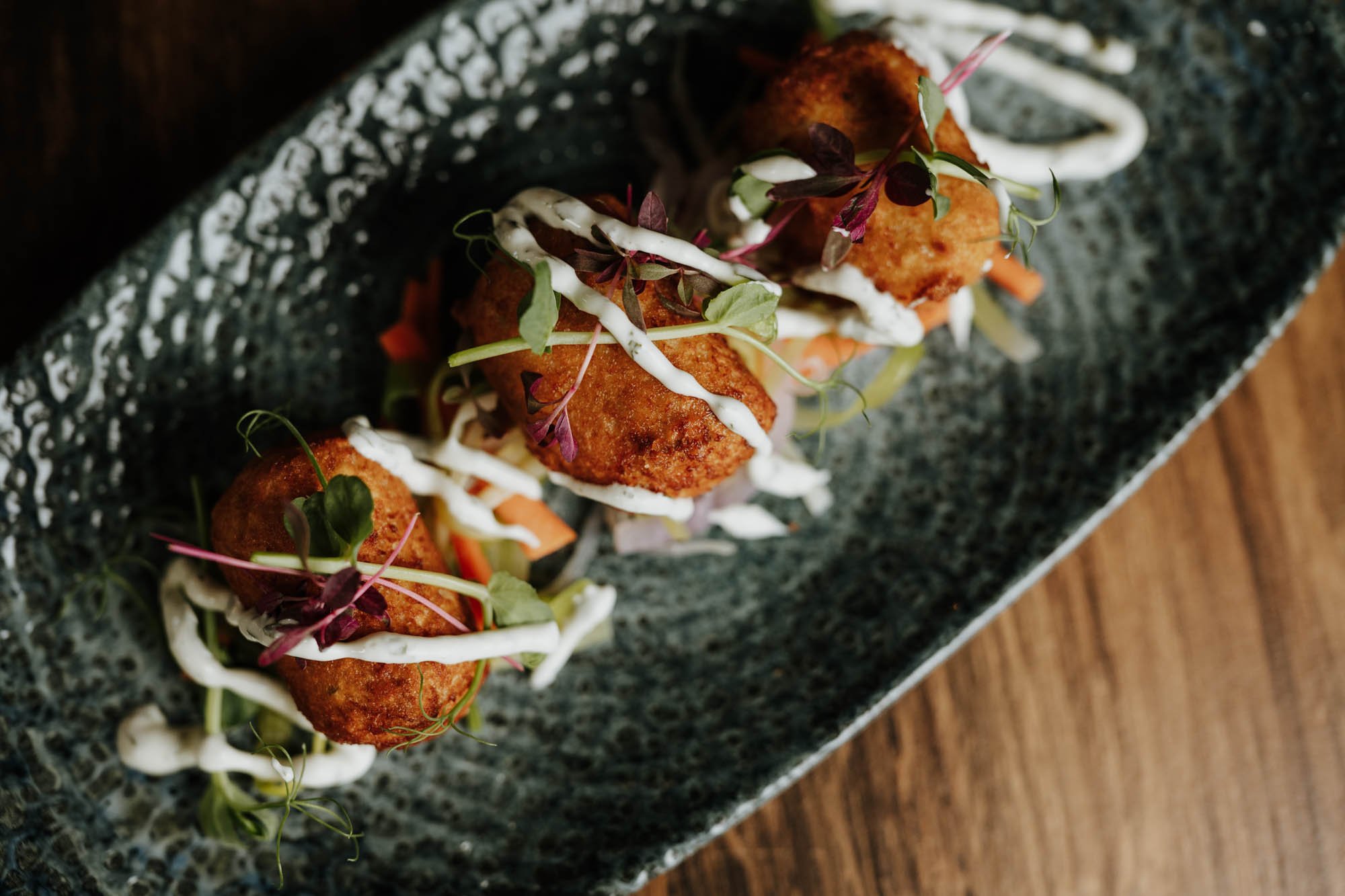 Three golden-brown fried croquettes garnished with microgreens and drizzled with white sauce, on a textured black rectangular serving plate.