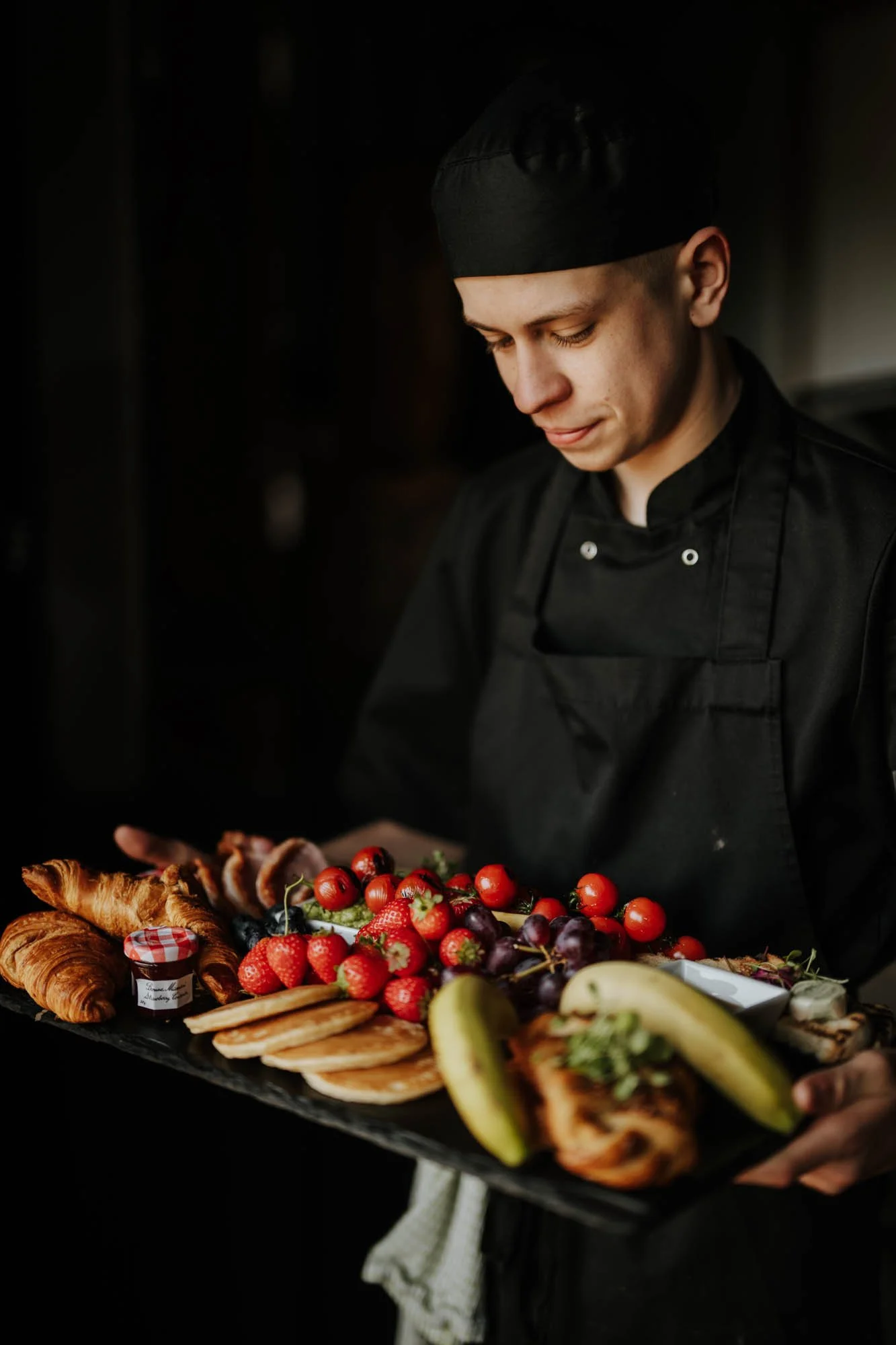Chef holding a tray of assorted pastries, berries, and fruits in a dark kitchen.