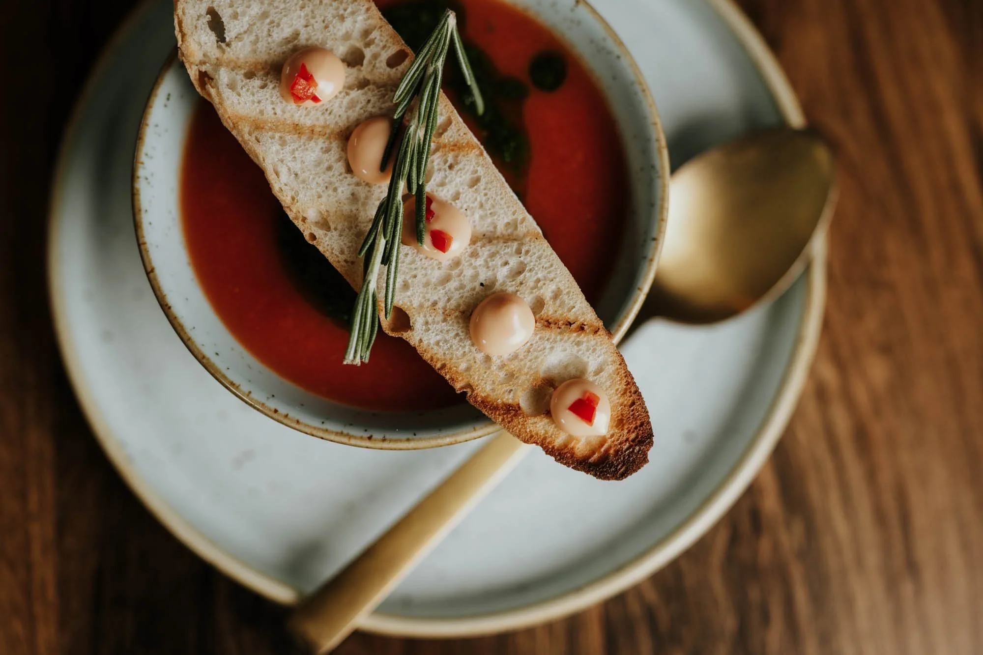 A bowl of tomato soup garnished with a slice of toasted bread topped with dollops of mayonnaise or cream and small red chili pieces, a sprig of rosemary for garnish, placed on a ceramic plate with a golden spoon resting on the plate.