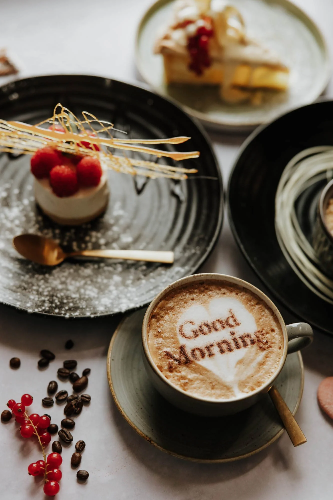 A cup of coffee with 'Good Morning' written in cocoa powder, a small dessert topped with raspberries, and plates of sliced cake and spaghetti on a white table.