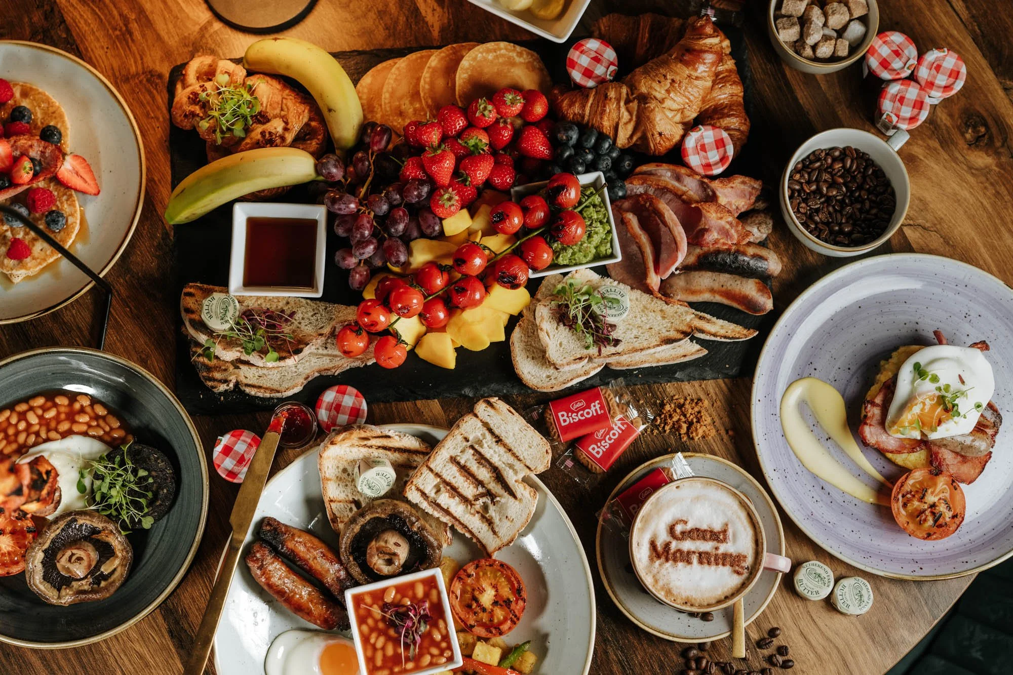 A breakfast spread with various foods including berries, croissants, toast, eggs, bacon, coffee, and syrup on a wooden table.
