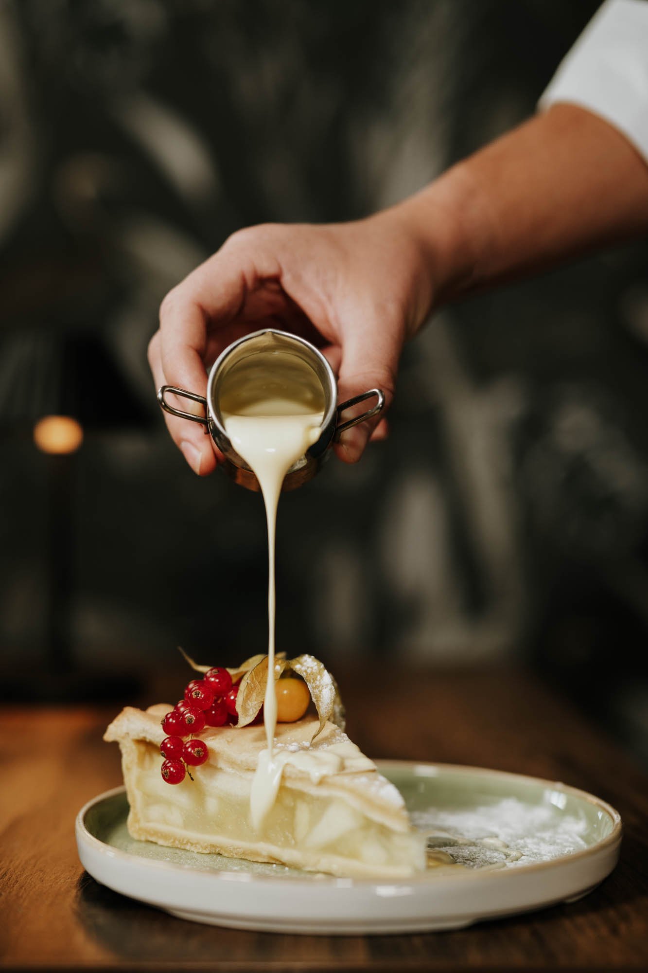 A person pouring white sauce over a slice of cream-filled pie garnished with red berries and a small golden fruit on a green plate.