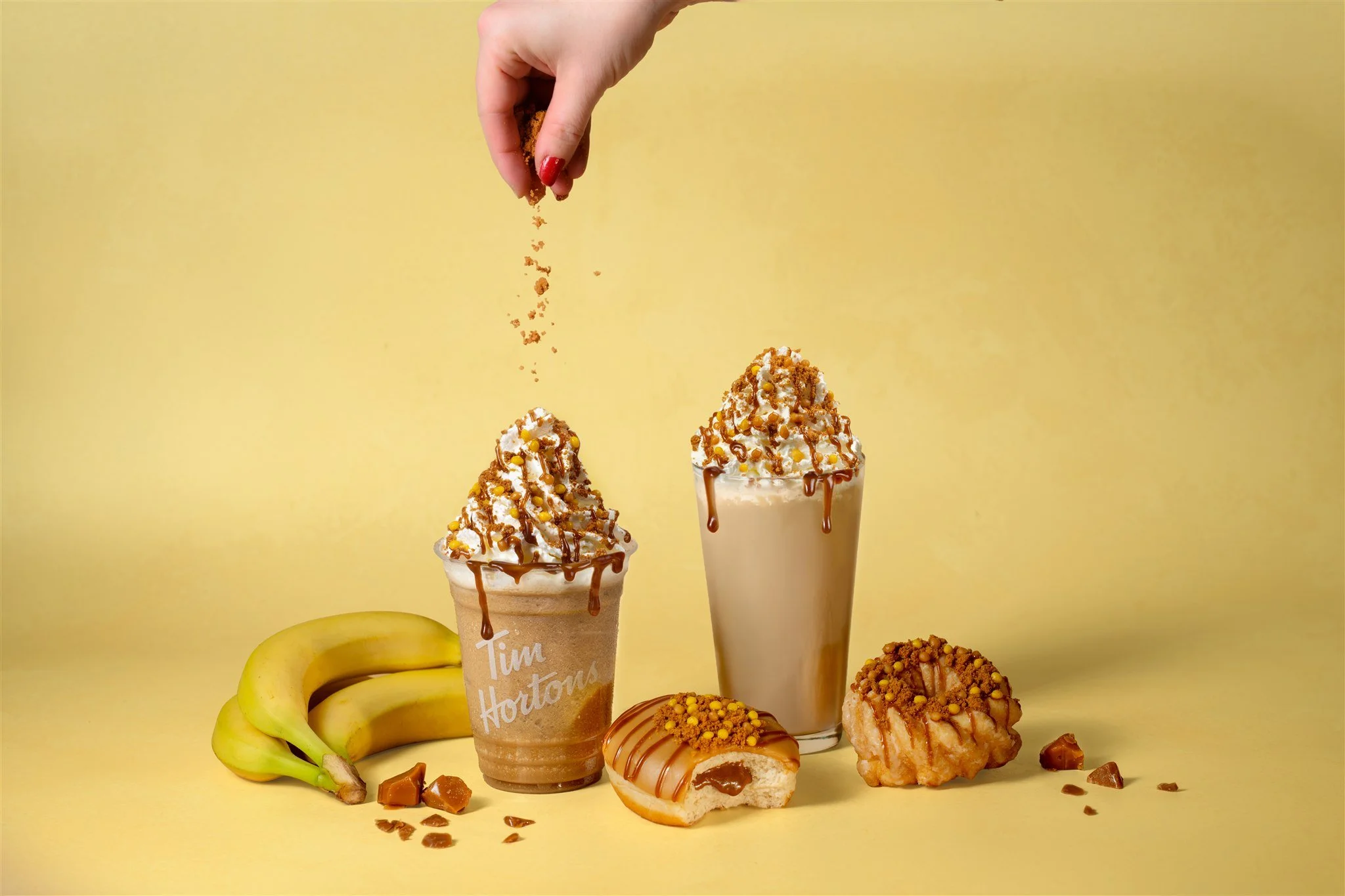 Two caramel-flavored iced drinks topped with whipped cream, caramel syrup, and sprinkled topping. Fresh bananas, caramel candies, a glazed donut, and a fried dessert are arranged on a yellow background.