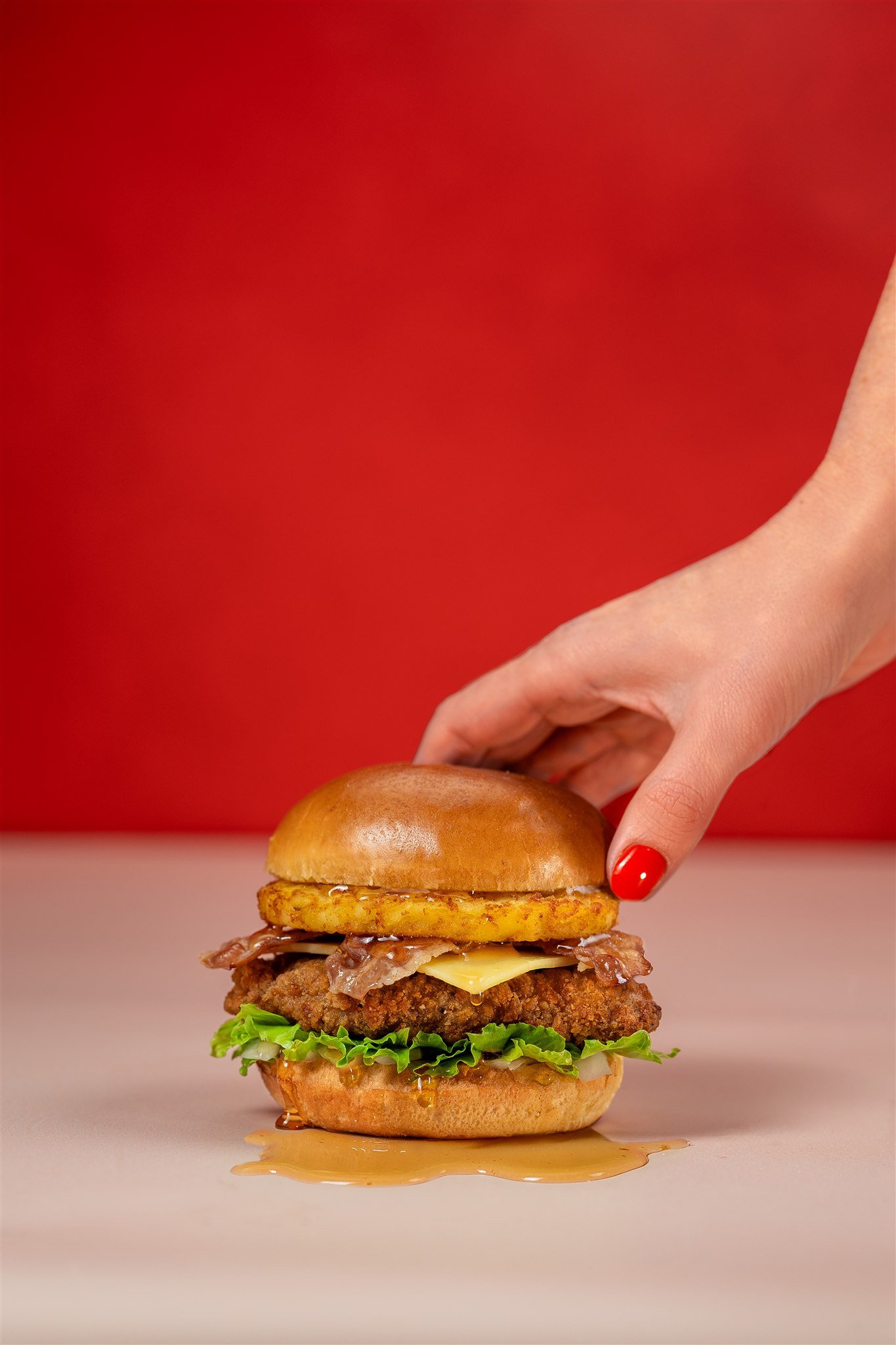 A person's hand with red nail polish holding a large hamburger with lettuce, fried chicken, cheese, bacon, a hash brown, and a brioche bun on a white surface with a red background.