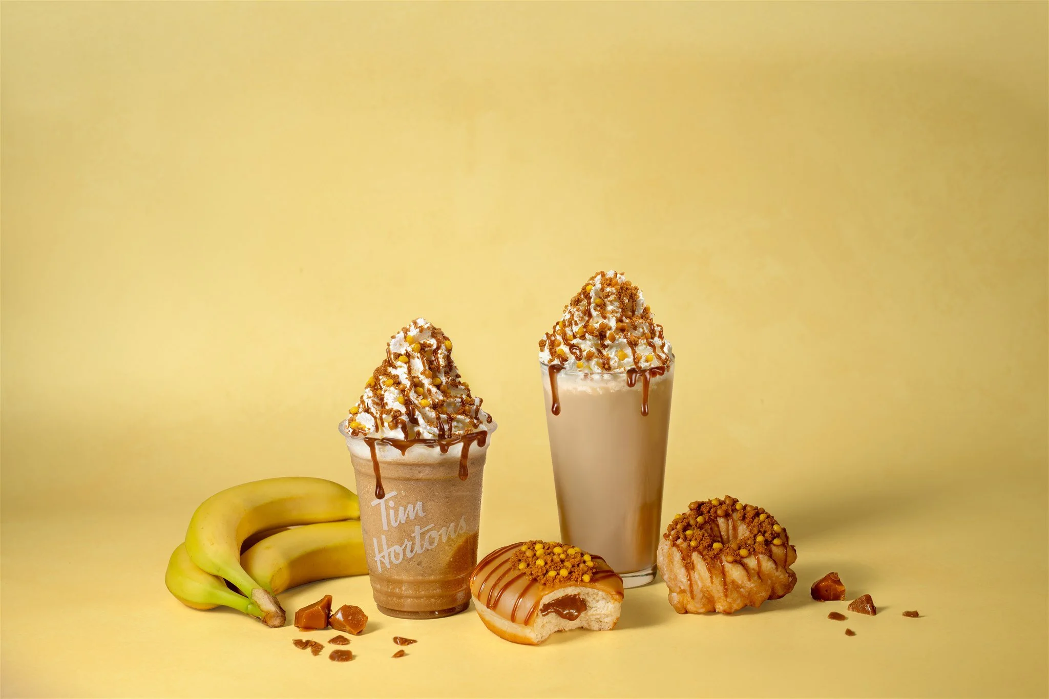 Two caramel-flavored drinks topped with whipped cream, caramel drizzle, and golden toppings, surrounded by bananas, caramel candies, and caramel-flavored baked goods on a yellow background.