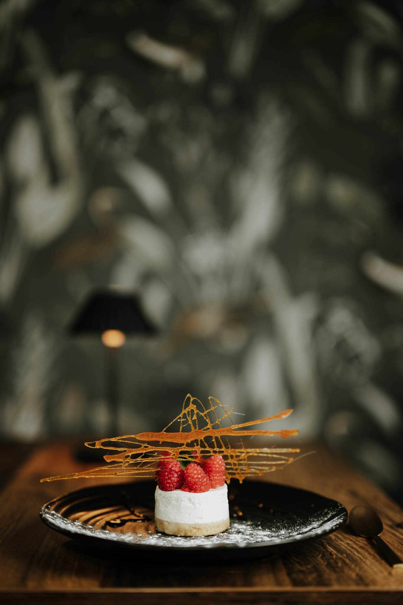 A dessert with raspberries, a white base, caramel lattice decoration, on a black plate with a spoon on a wooden table.