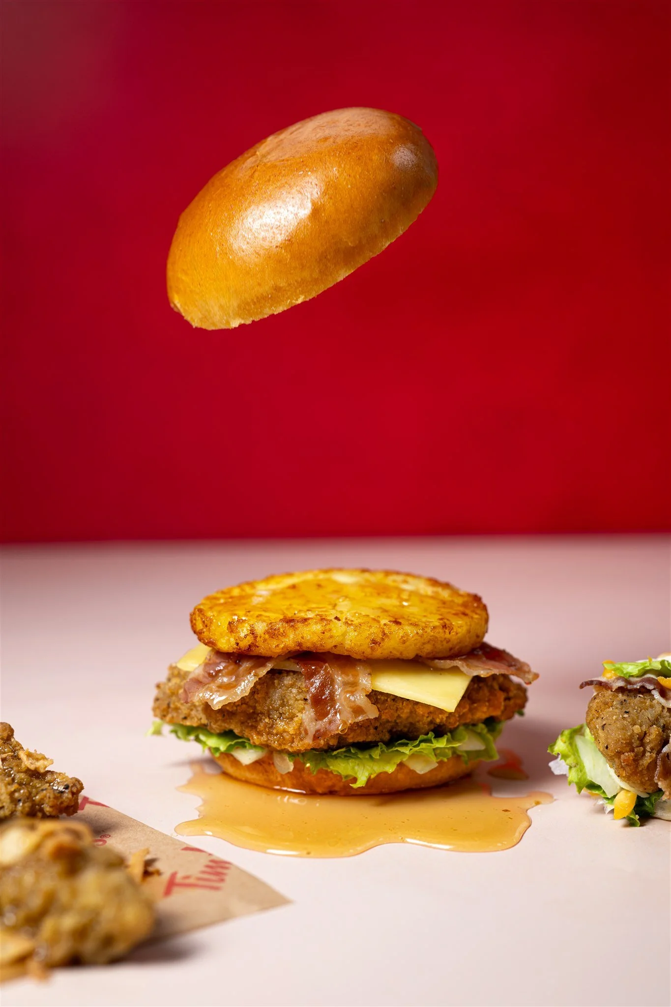 A chicken sandwich with lettuce, cheese, bacon, a fried chicken patty, and a hash brown on top, with a falling biscuit overhead, and spills of gravy on a white surface against a red background.