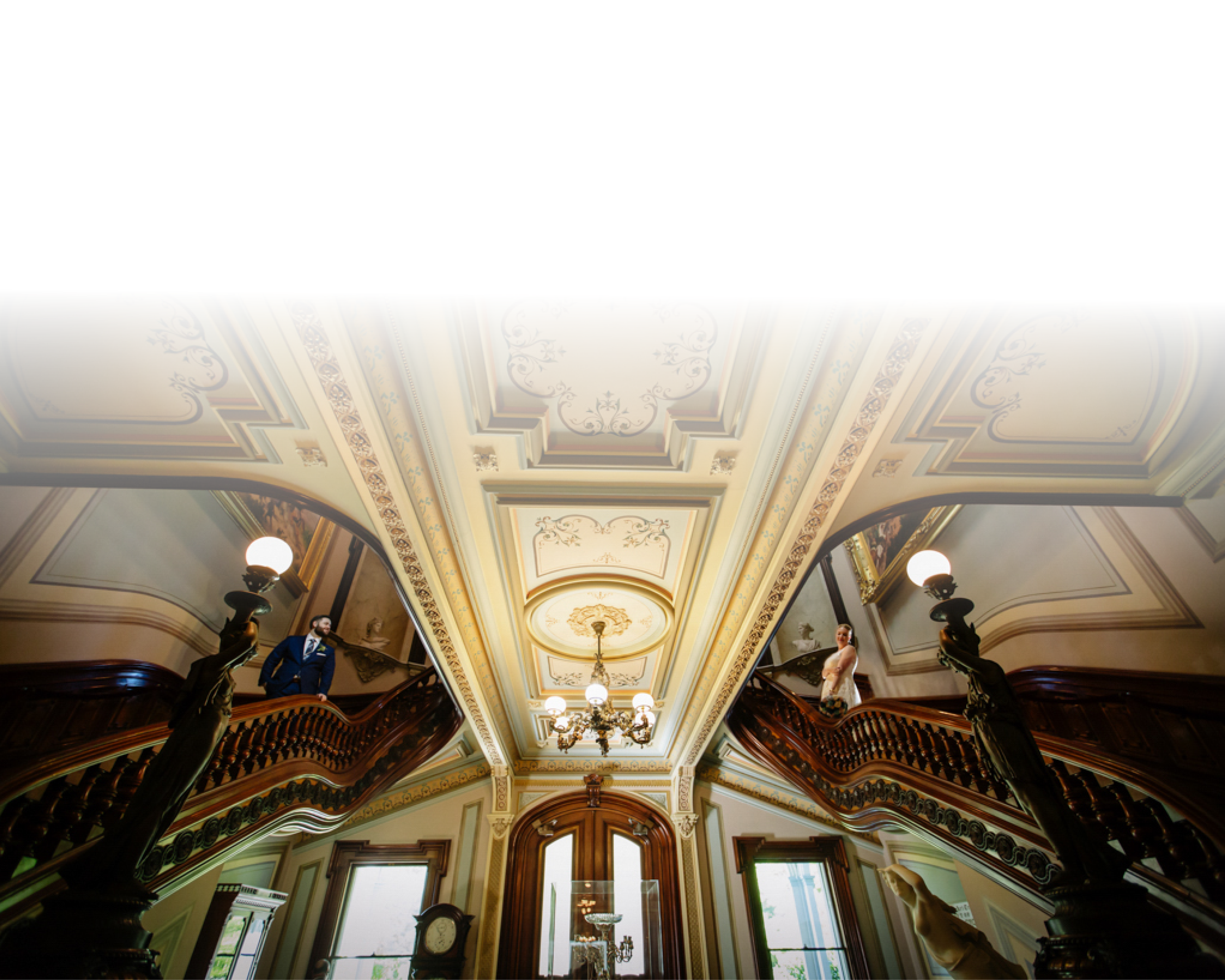 Interior of a grand, ornate mansion with a high, decorated ceiling, chandelier, staircase, and two elegantly dressed people standing on the staircase balustrades.