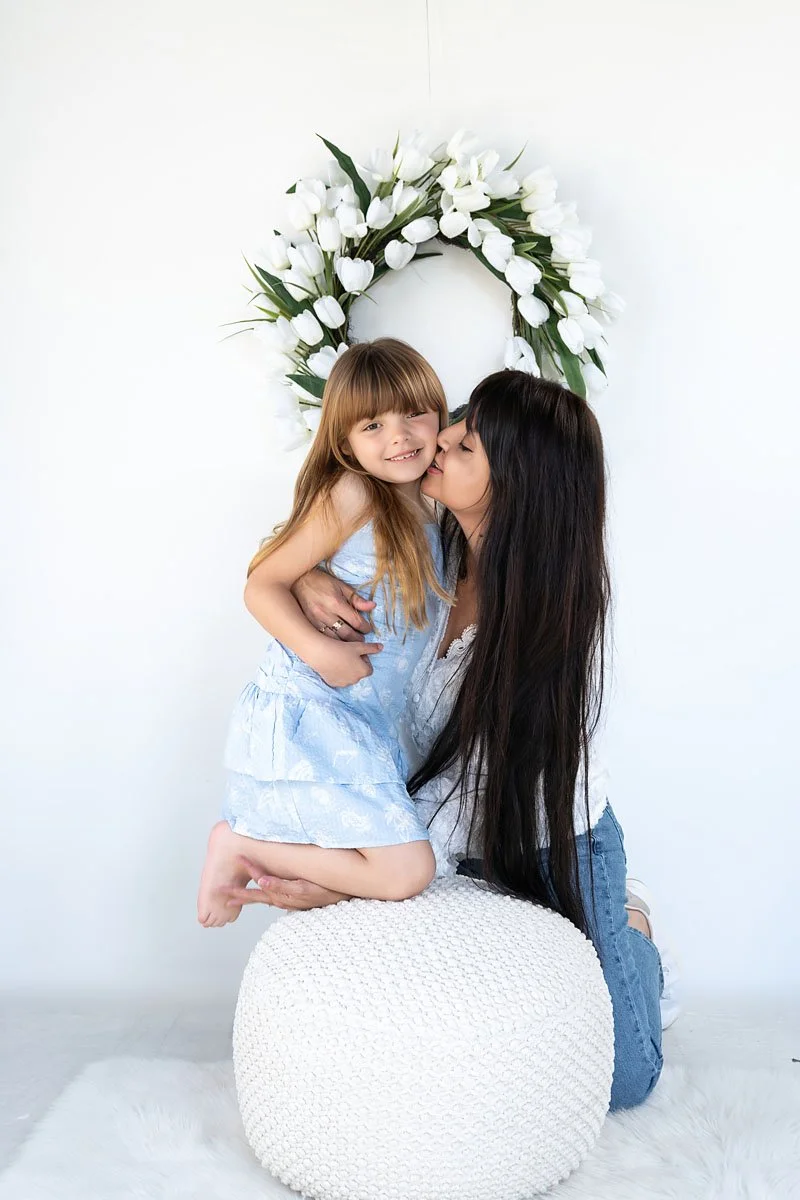 A woman kneeling and holding a young girl, who is sitting on her lap, in front of a white wall with a floral wreath above. The woman is kissing the girl on the cheek, and the girl is smiling at the camera. The scene has a soft, cozy setting with a white pouf and furry rug.