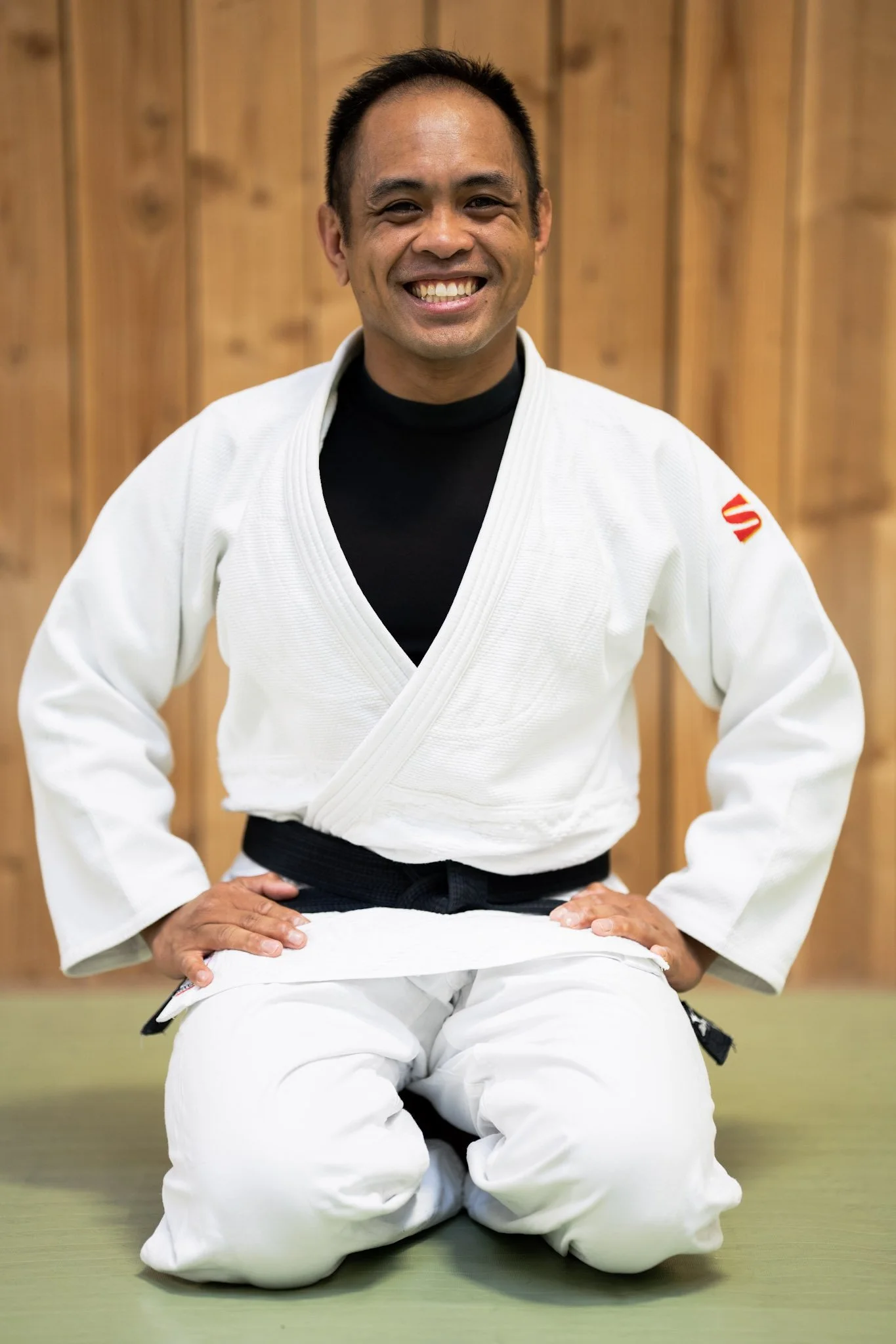 A smiling man in a white martial arts gi with Japanese characters on the chest, standing in front of a wooden wall.