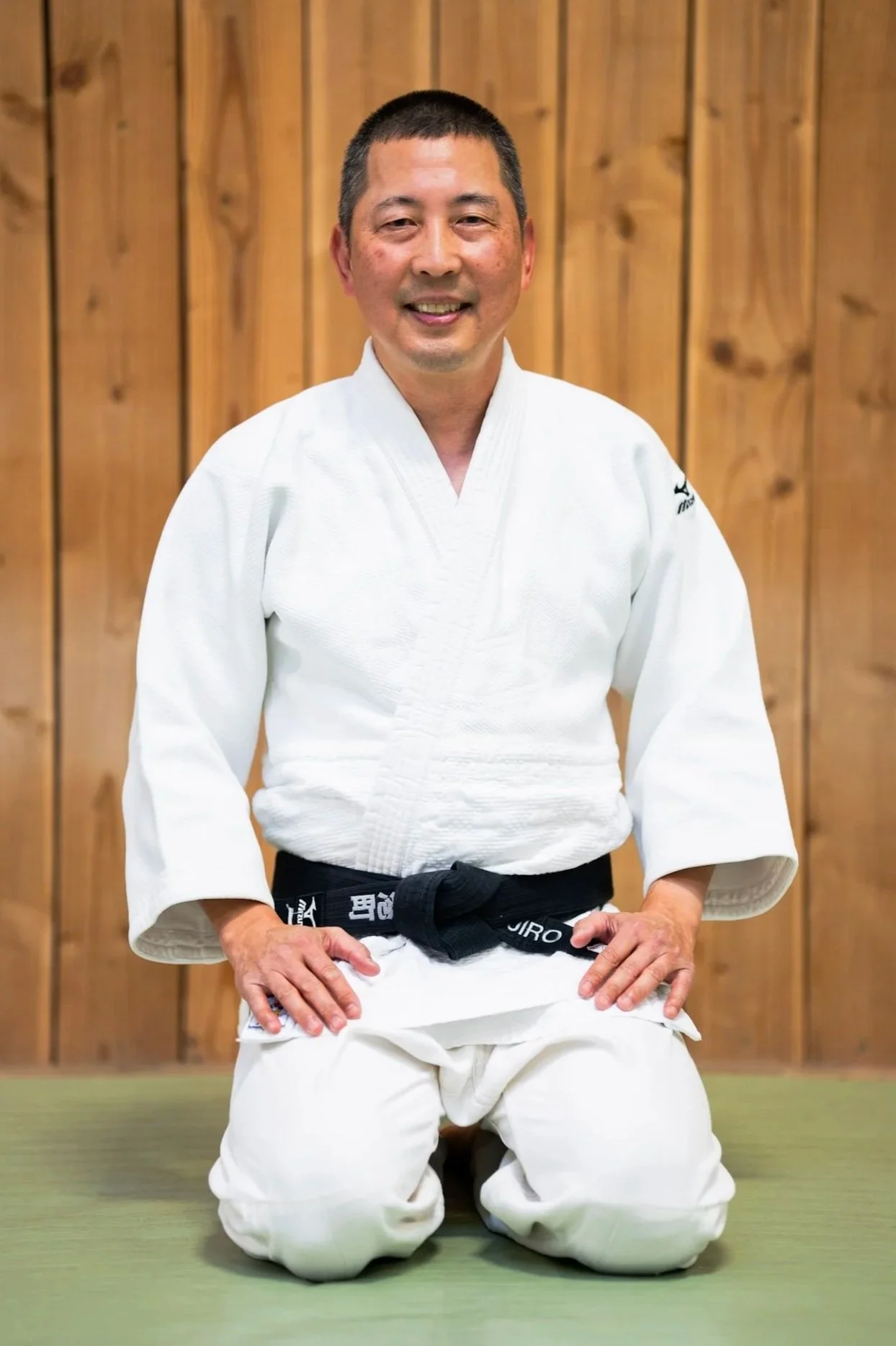 A man smiling in a white judo gi with a black belt standing in front of a wooden wall.