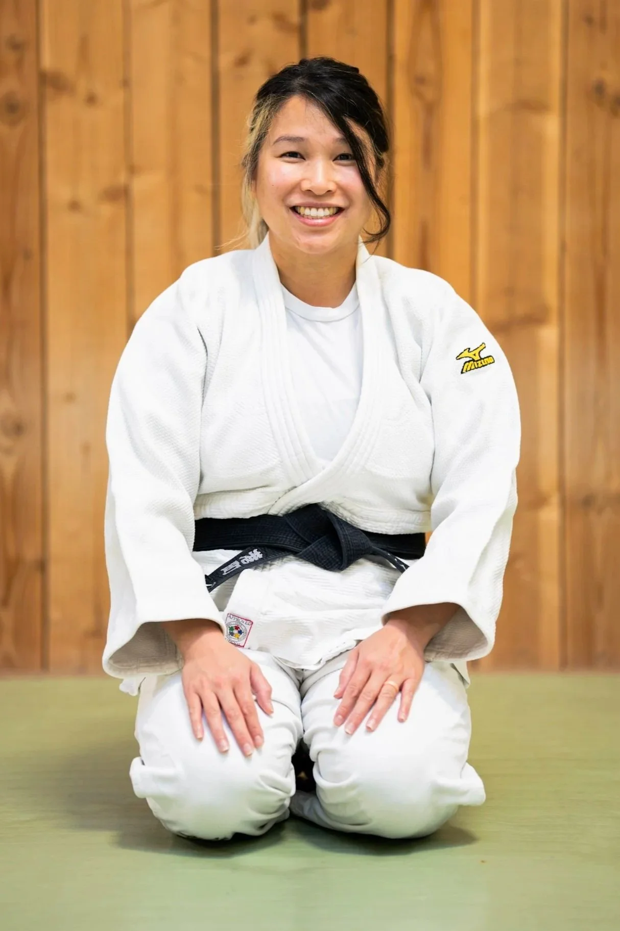 A smiling young woman wearing a white judo gi and a black belt, standing in front of a wooden wall.