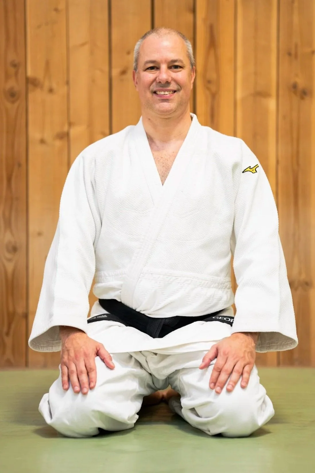 A man standing in a martial arts dojo or gym, wearing a white judo gi with a black belt that has his name, George, embroidered on it. He is smiling and standing in front of a wooden wall with a fabric ceiling above.