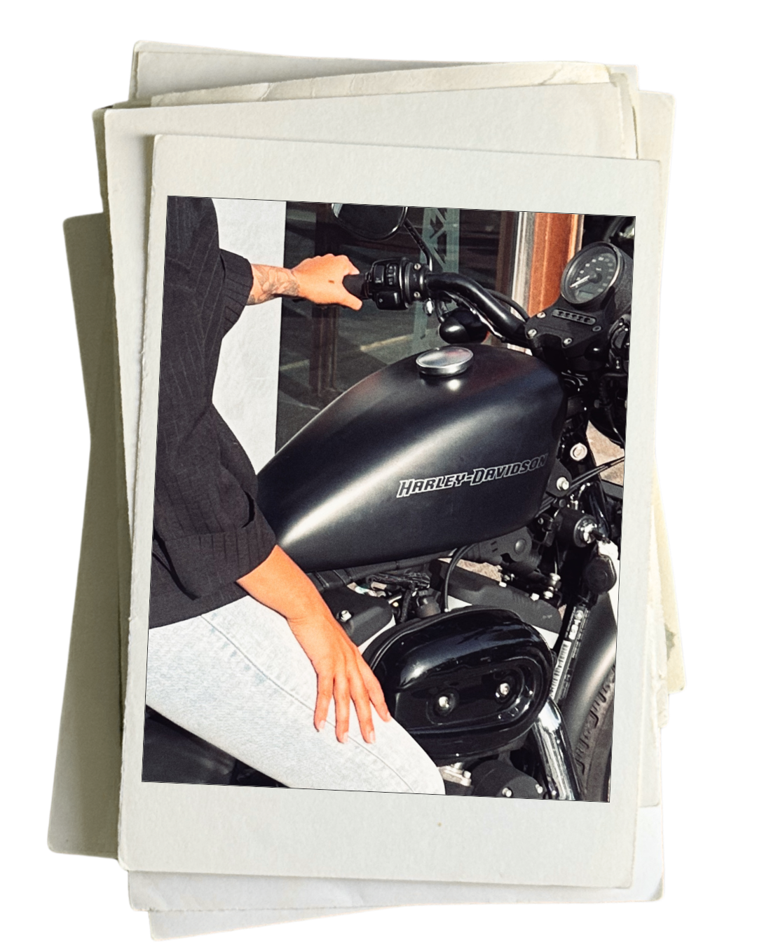 A person sitting on a black Harley-Davidson motorcycle, wearing a black sweater and gray sweatpants, with their left hand resting on their thigh and right hand on the handlebar.