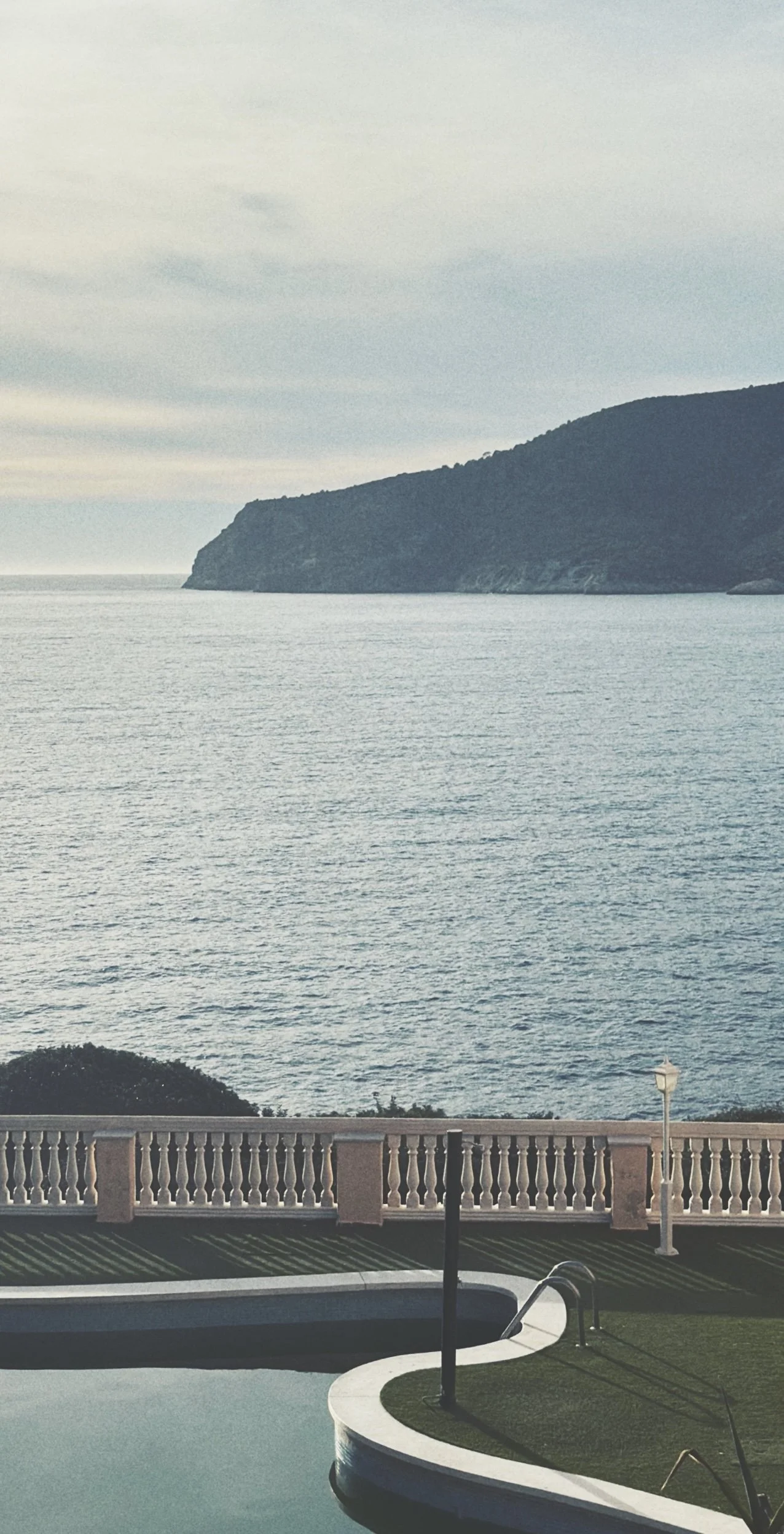 Coastal view with water, a headland, cloudy sky, and a pool area with a fence and lamp post.