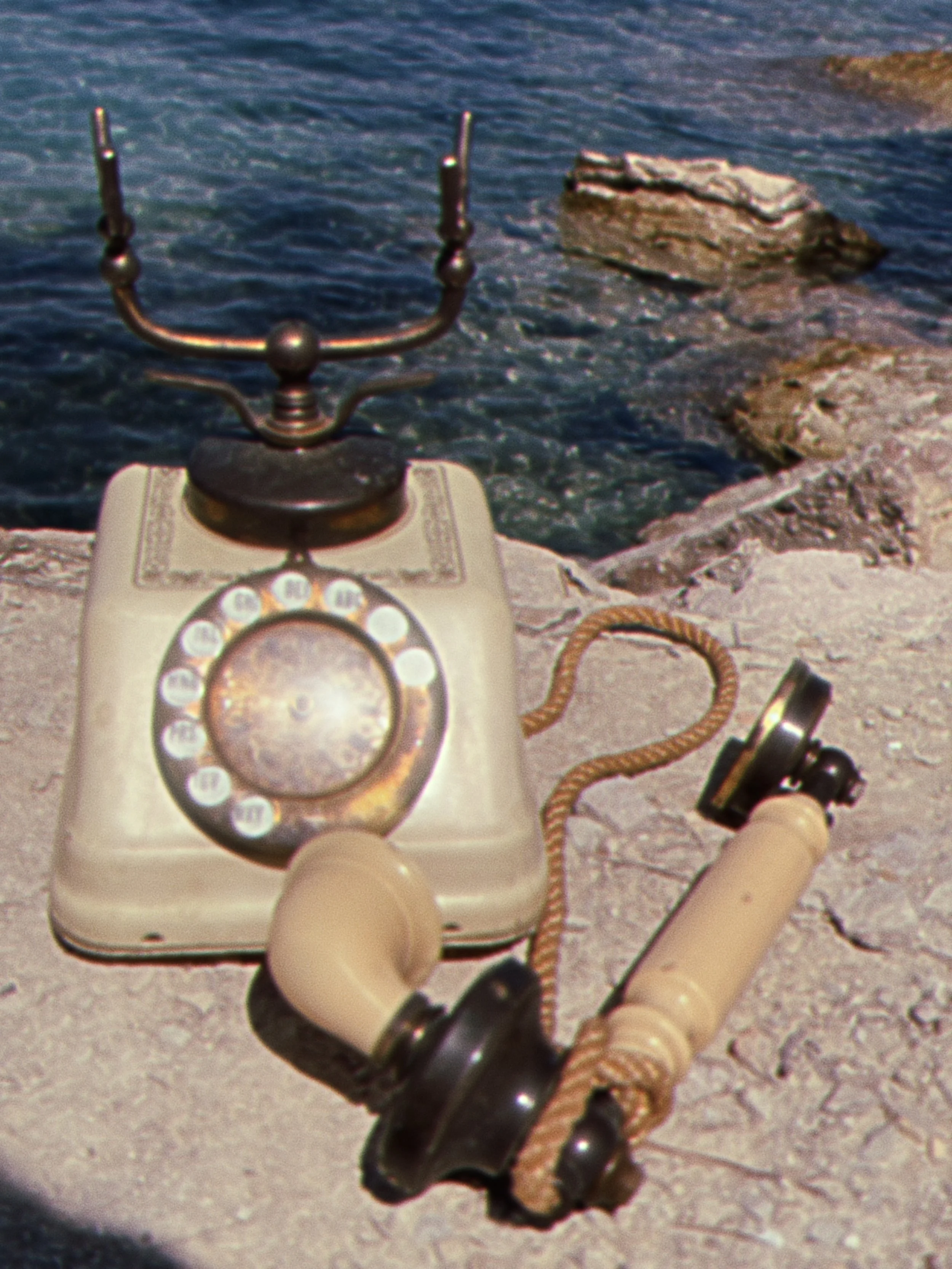 A vintage rotary telephone is on a table, with a set of beige earbuds with black ends held in a person's hand in the foreground, near the phone. In the background, there is a body of water with rocks along the shore.