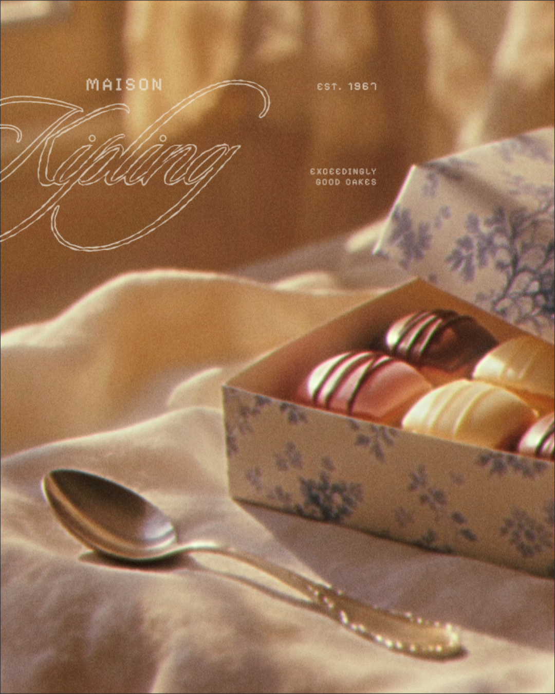 A box of assorted macarons with pink, yellow, and brown colors, decorated with chocolate drizzle, resting on a cream-colored cloth beside a silver spoon.