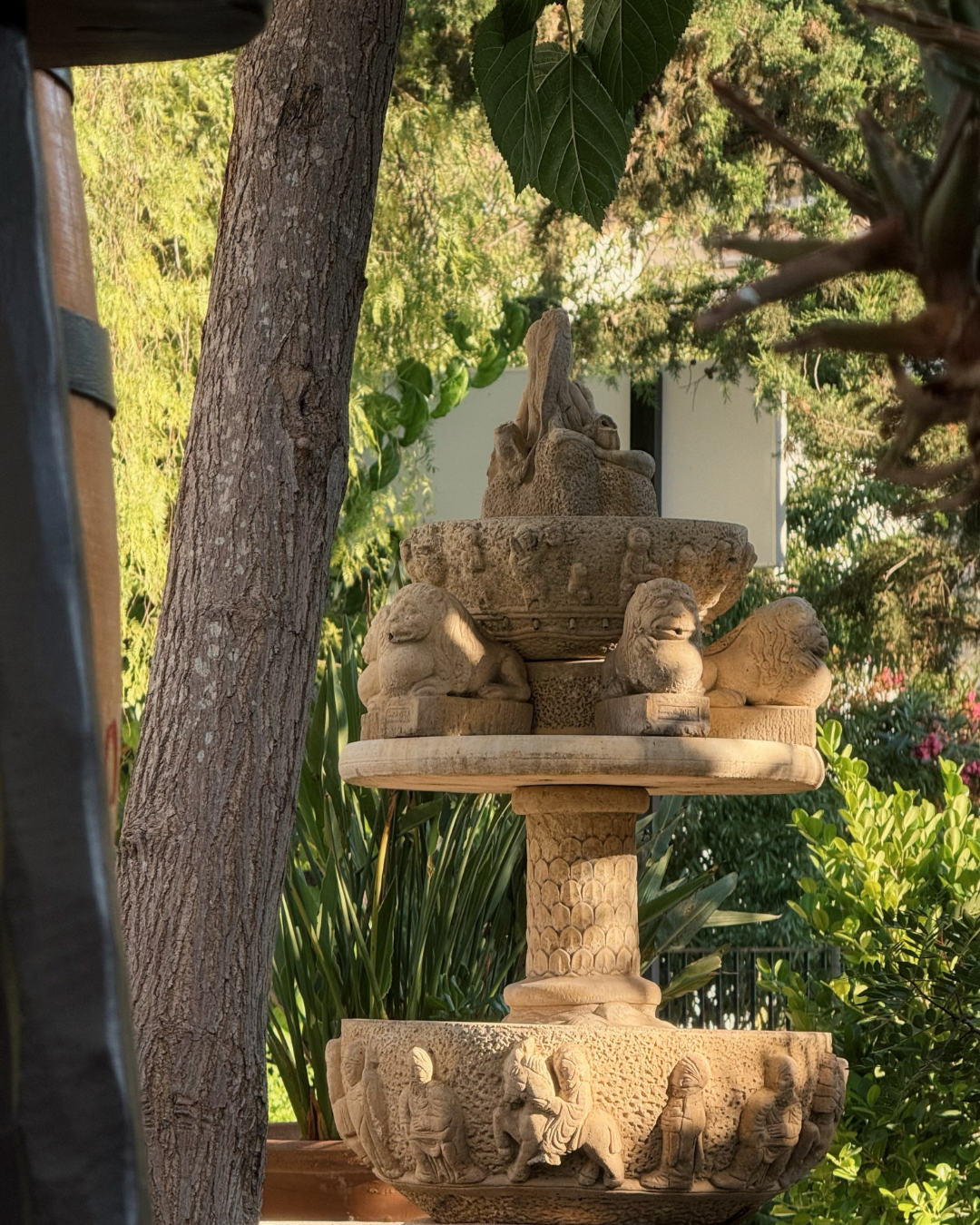 Stone sculpture fountain with carved animals and mythological figures, surrounded by lush green plants and trees.