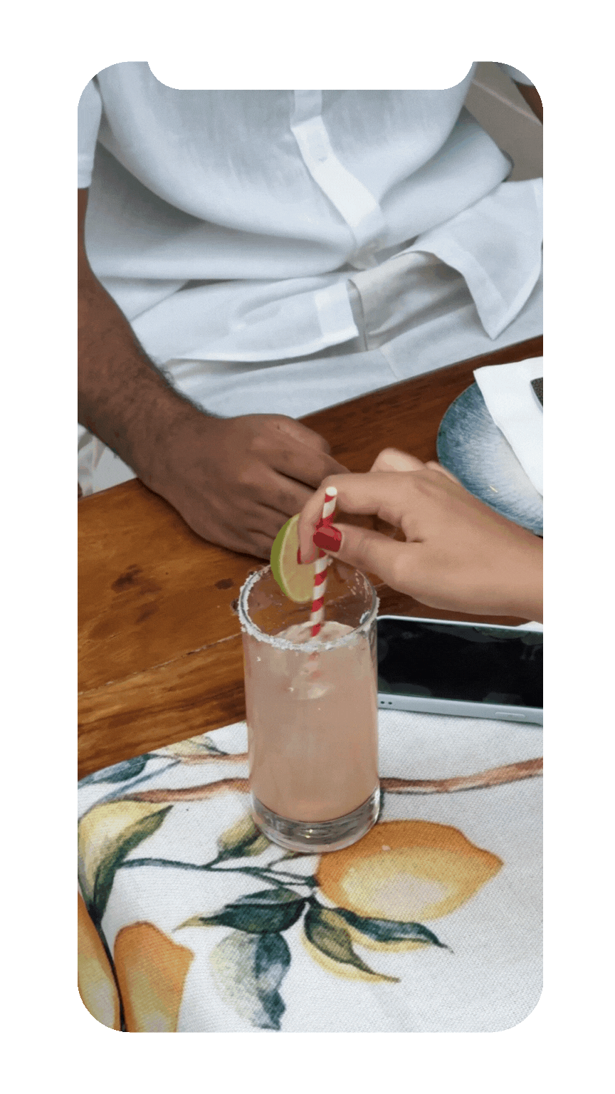A person in a white shirt offers a pink frozen drink garnished with a lime wedge, a salted rim, and a red-and-white striped straw to another person with red-painted nails at a table with a lemon-themed tablecloth.