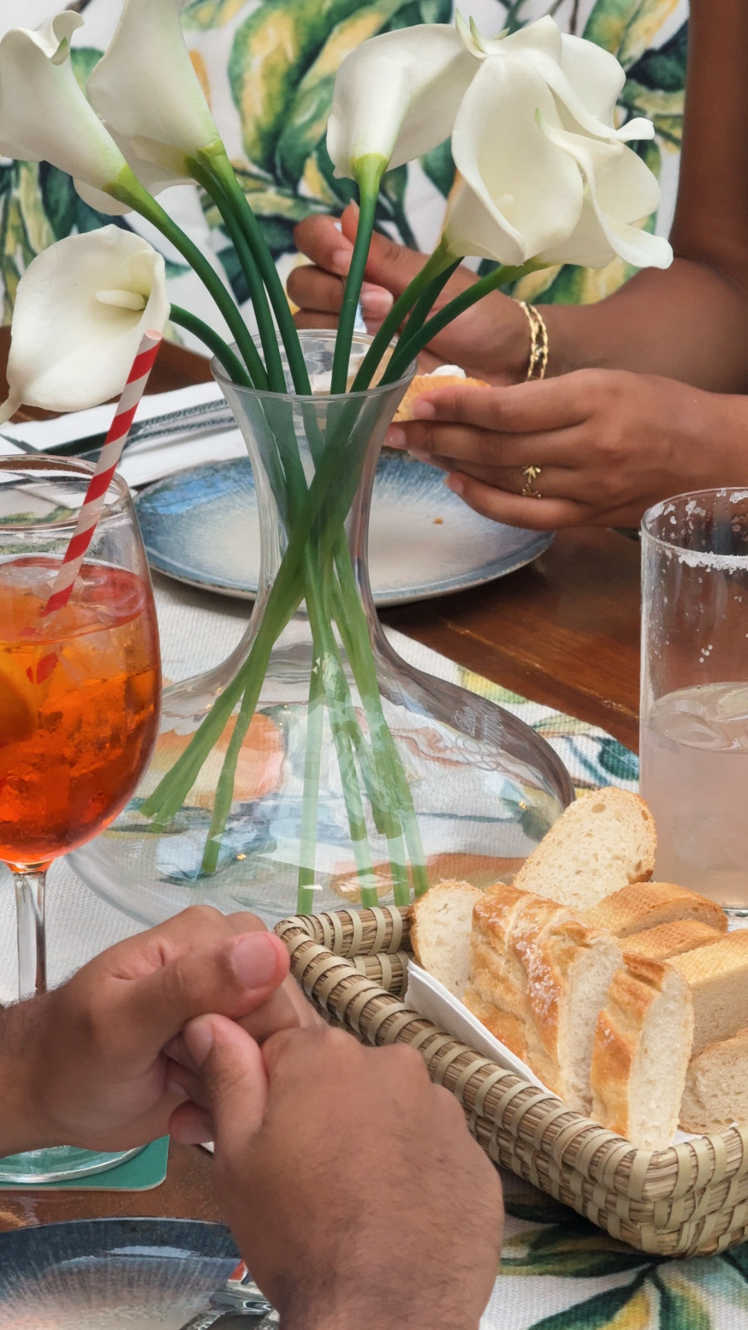 A dining table with a bouquet of white calla lilies in a glass vase, a plate with bread, a glass of iced tea with a straw, and a glass of ice water. People are holding hands.