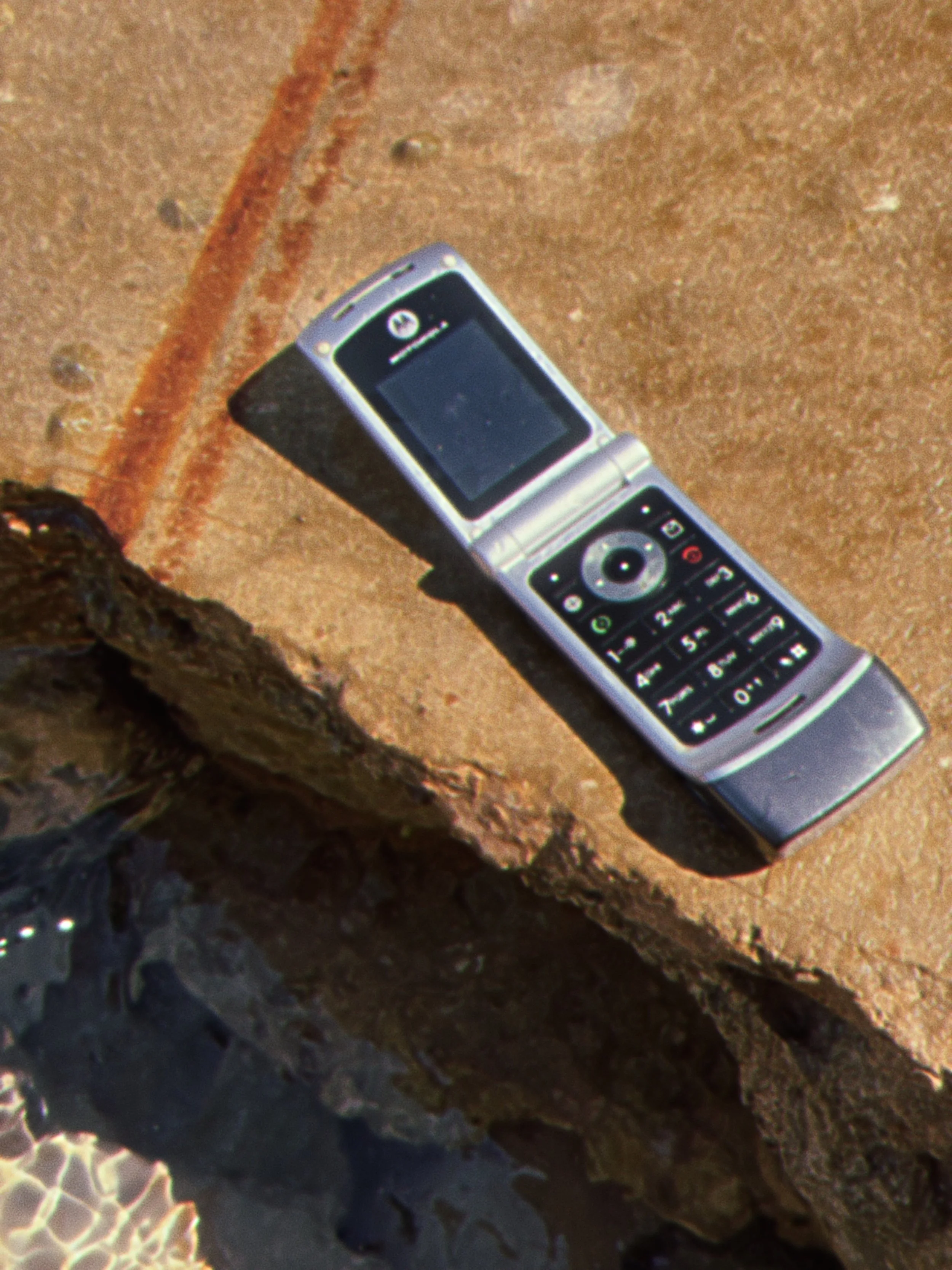 A flip phone with a colorful keypad resting on a sandy surface near seaweed and coral.
