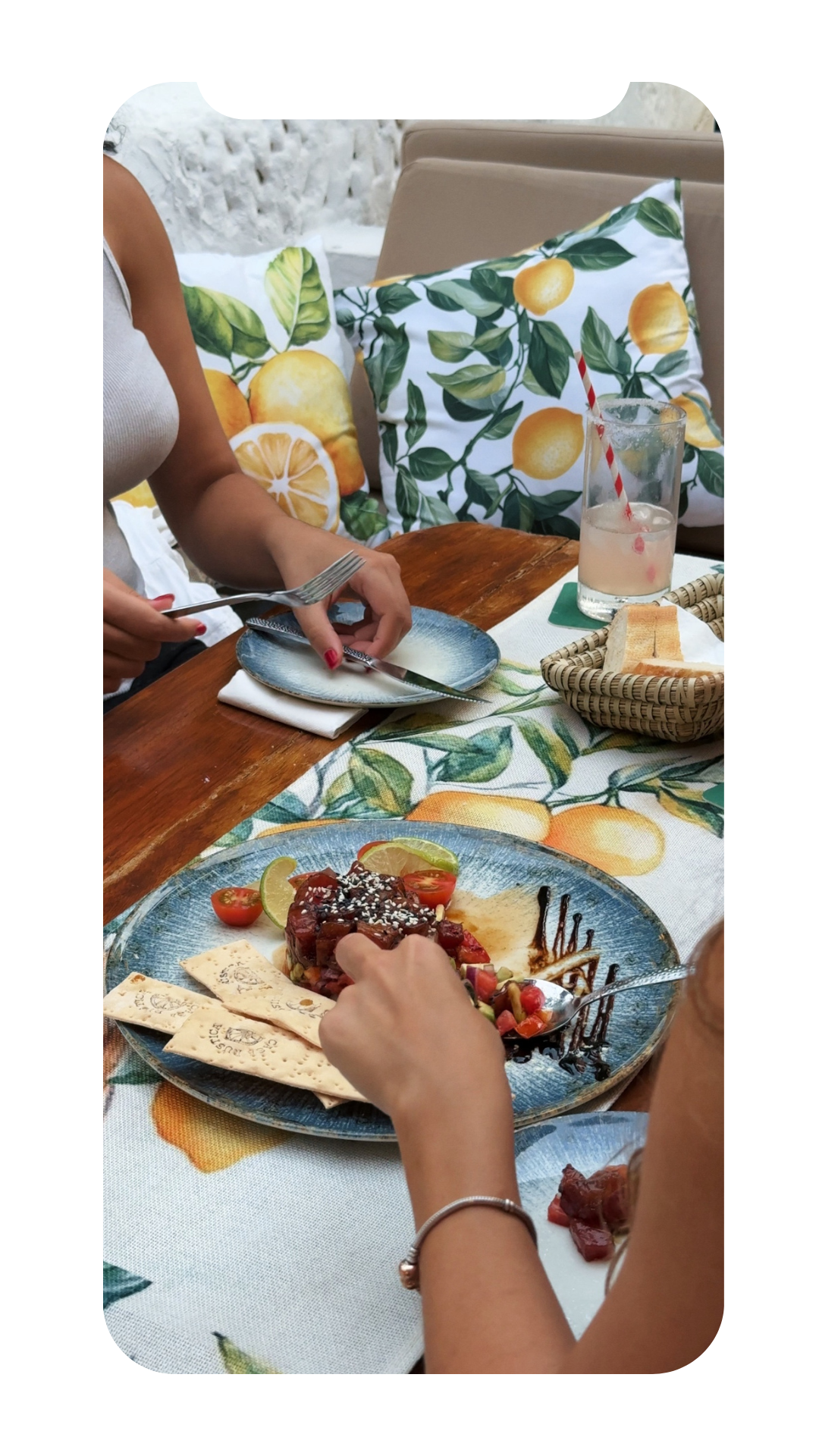 People dining at a restaurant table with a plate of dessert, slices of lime and cherry tomatoes, and a glass of lemonade on a lemon-themed tablecloth.