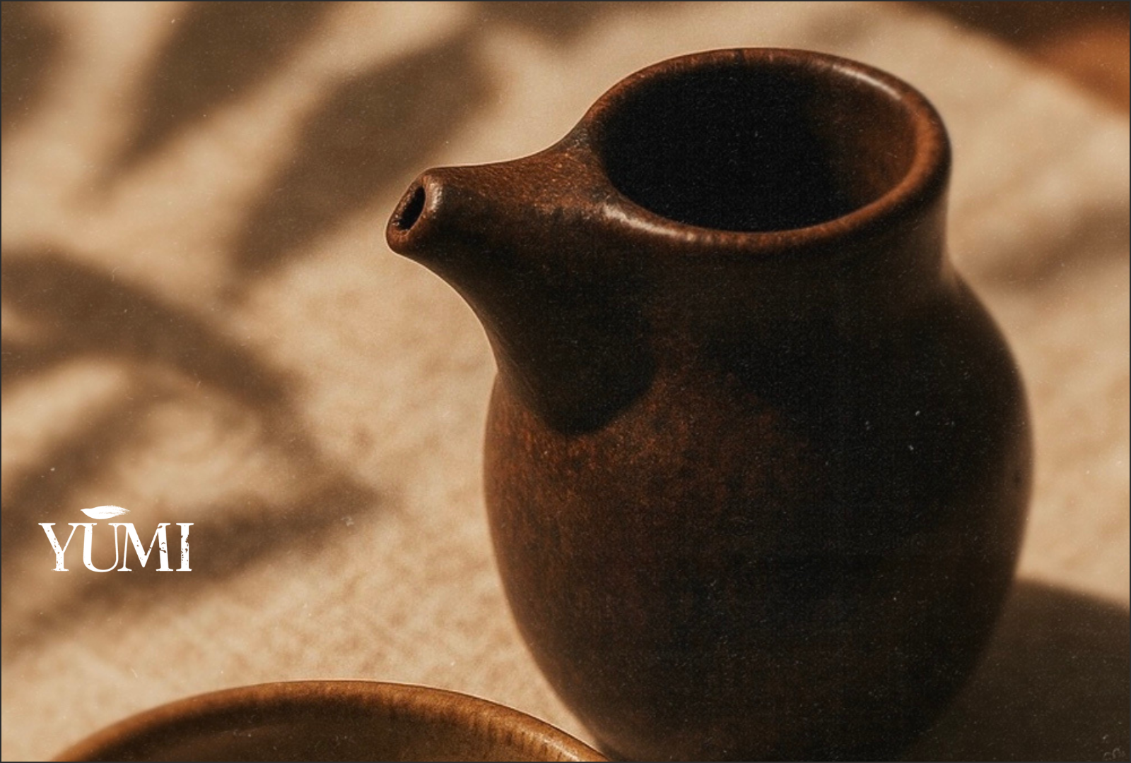 Close-up of a small, dark brown ceramic teapot with a rounded body and a short spout, placed on a light beige textured surface, with the word 'YUMI' in white text at the bottom left corner.