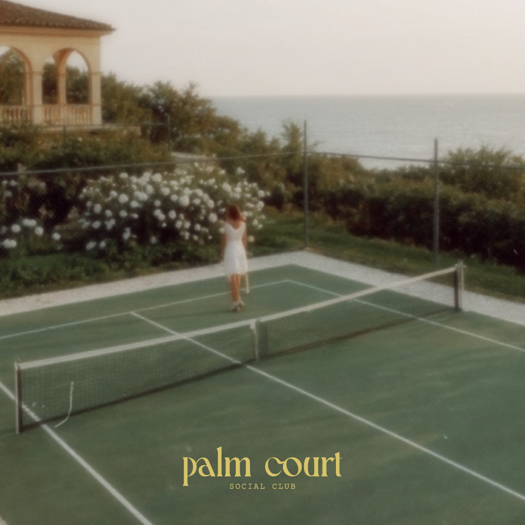 A woman in a white dress standing on a tennis court near The ocean, with a house and flowering bushes nearby, at sunset, with the text 'palm court social club' at the bottom.
