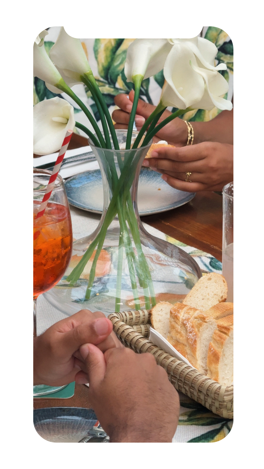 A dining table with a glass vase holding white calla lilies, a basket of sliced bread, a glass of iced tea with a red-and-white striped straw, and people's hands clasped together in the foreground.