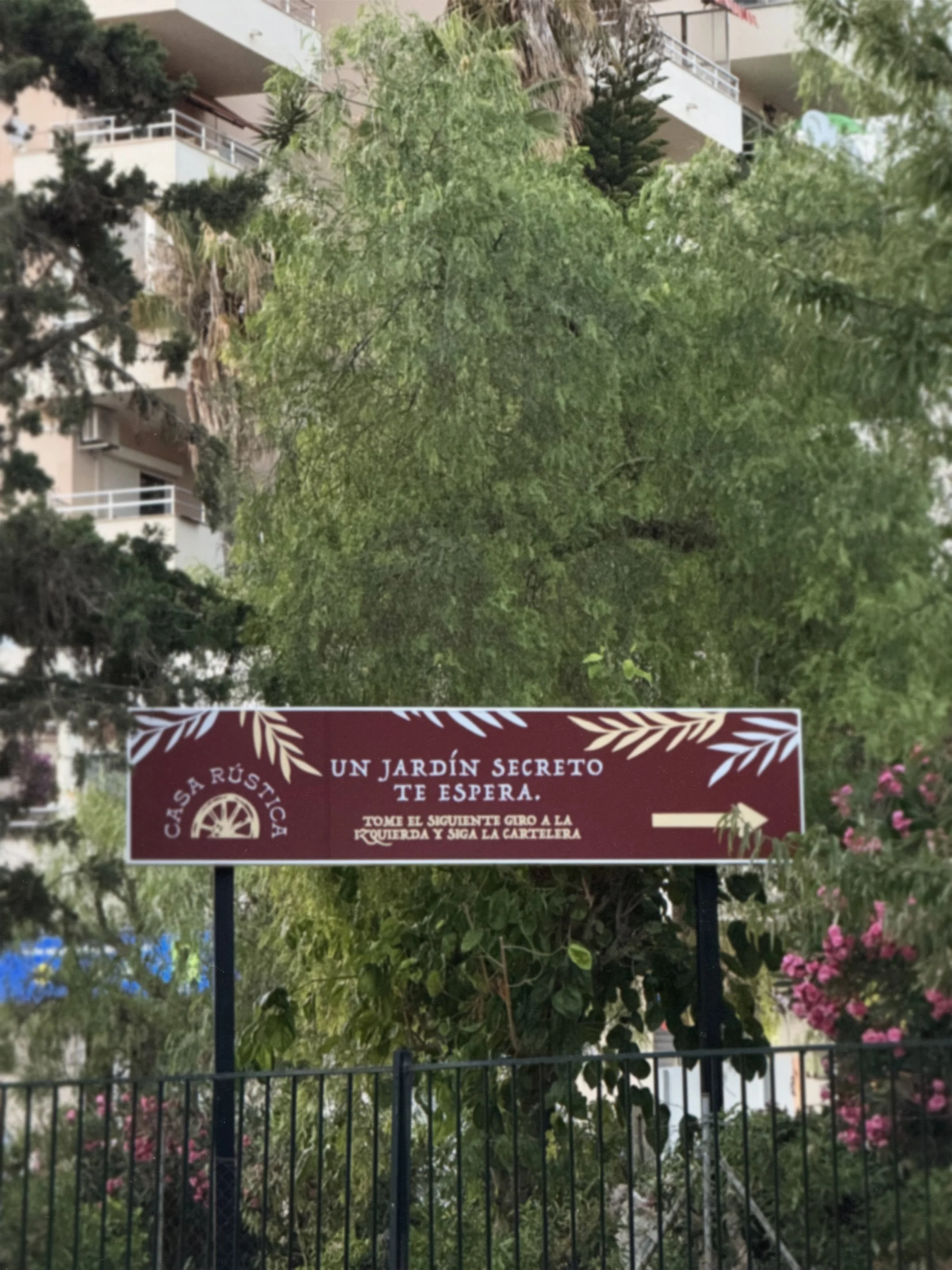 A sign that reads: "Casa Rústica, Un jardín secreto te espera. Toma el siguiente giro a la izquierda y sigue la cartelera" in front of a garden with trees and pink flowers, with buildings in the background.