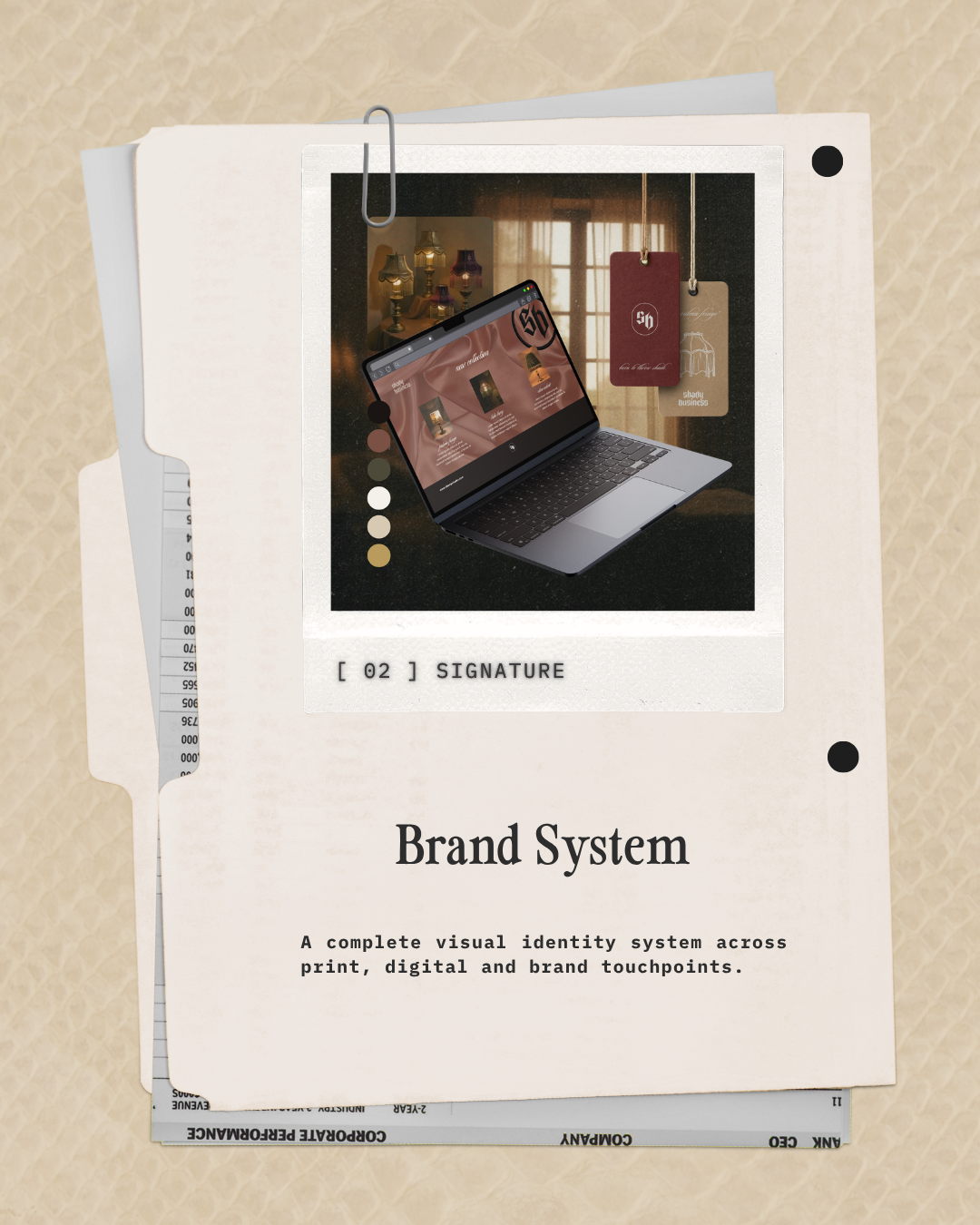 A printed branding guide titled 'Brand System' featuring a photo of a laptop displaying a website, with hanging tags and additional printed documents arranged on a beige background
