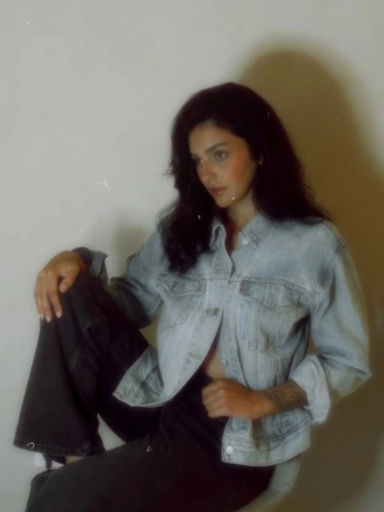 A woman with dark hair, wearing a jean jacket and black pants, sitting against a neutral wall, holding a black jacket