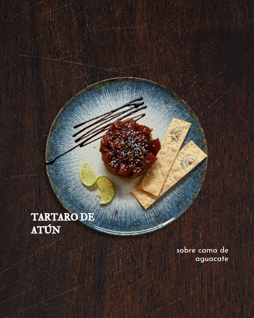 Tuna tartare on a bed of avocado, garnished with sesame seeds, served with lime wedges, accompanied by two saltine crackers, and drizzled with balsamic vinegar on a blue ceramic plate placed on a wooden surface.