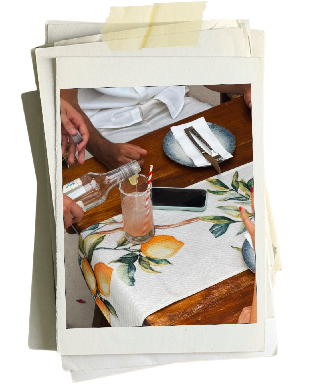 A close-up of a table setting with a floral tablecloth, a black phone, a plate with a napkin and fork, and a person pouring a drink with a straw and a slice of lime at a restaurant.