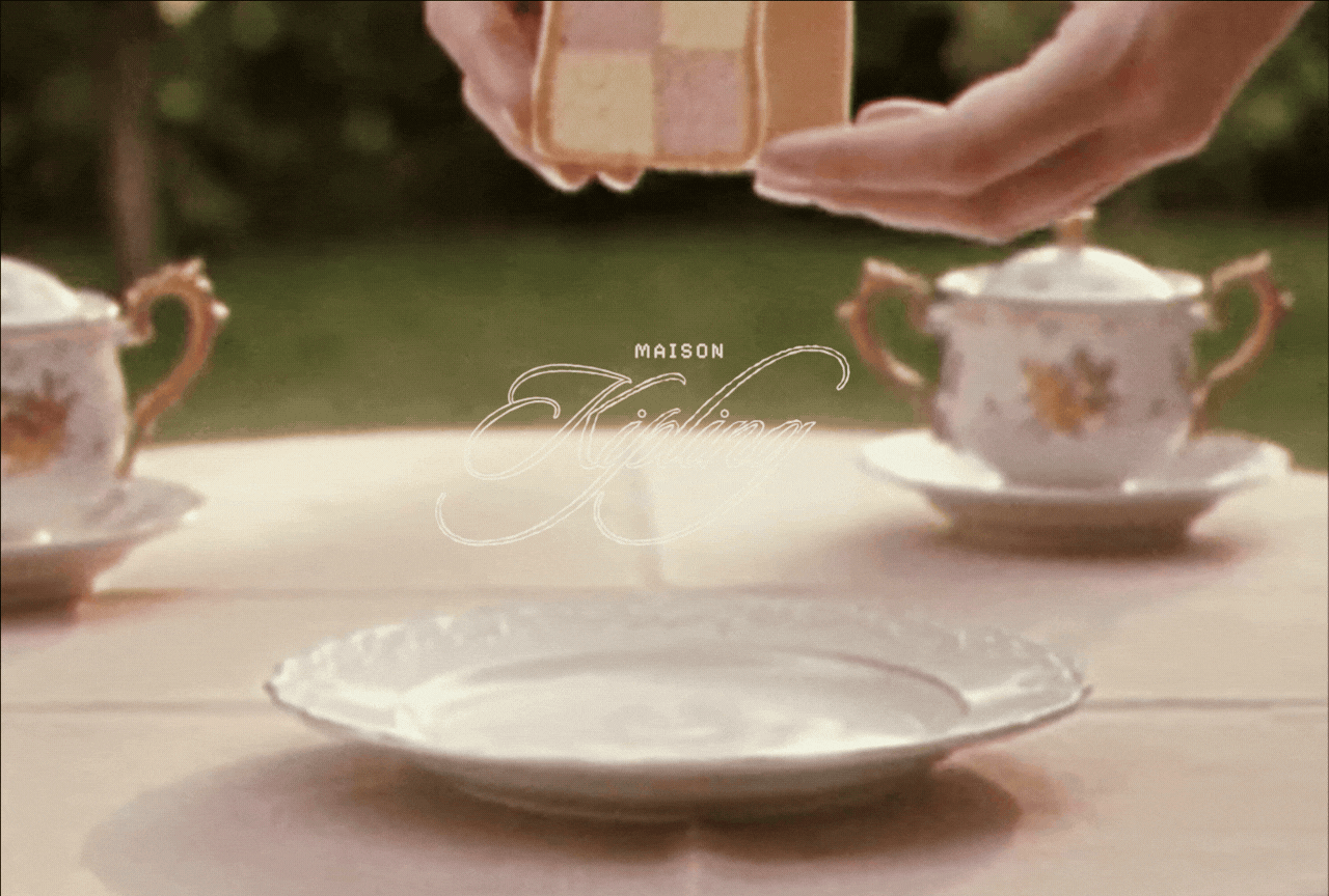 A tea set with two ornate teacups and a teapot on a table, with a person's hand holding a bowl or small container above the table. There are trees in the background.
