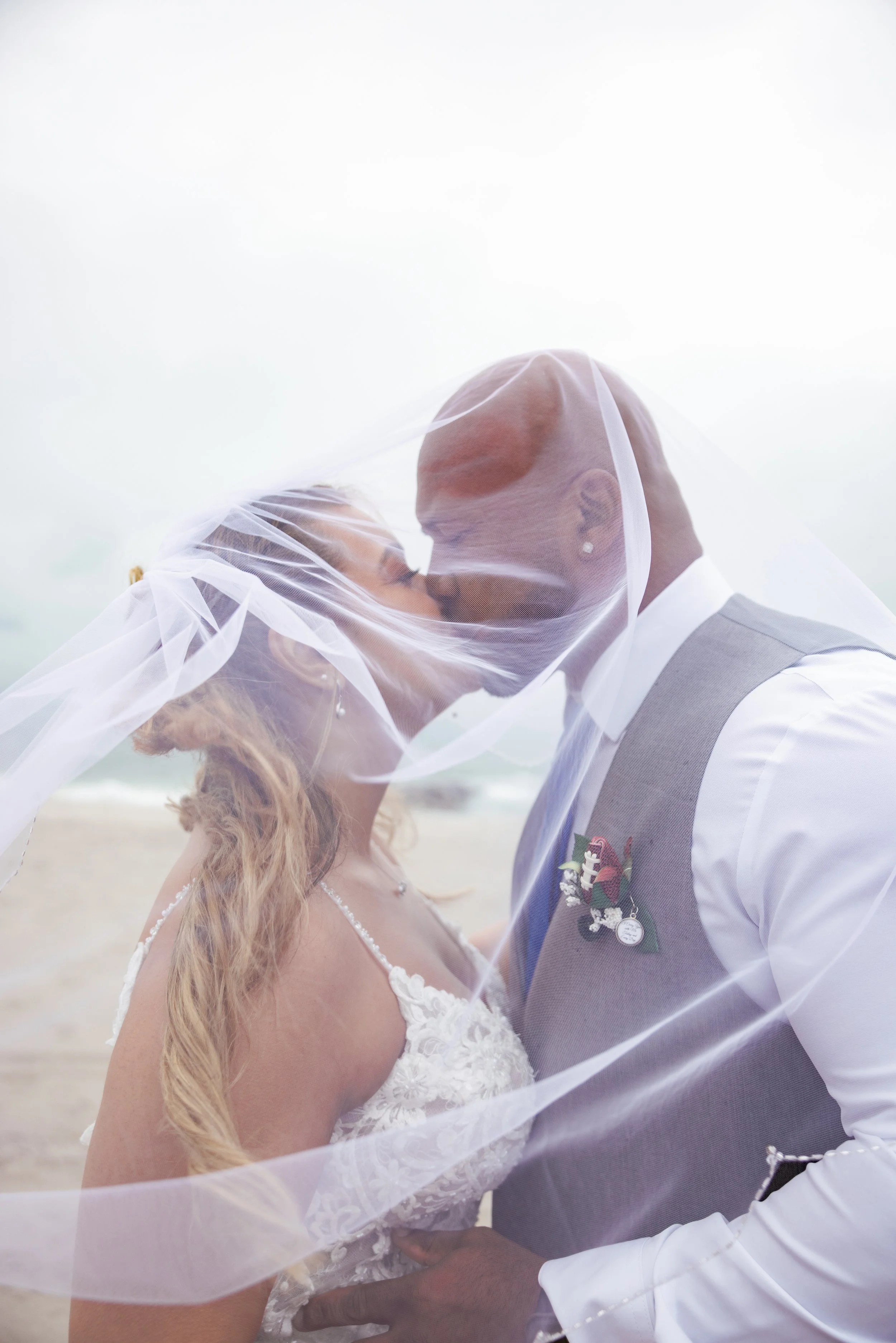 A bride and groom kissing on the beach, with their faces partially obscured by a veil.