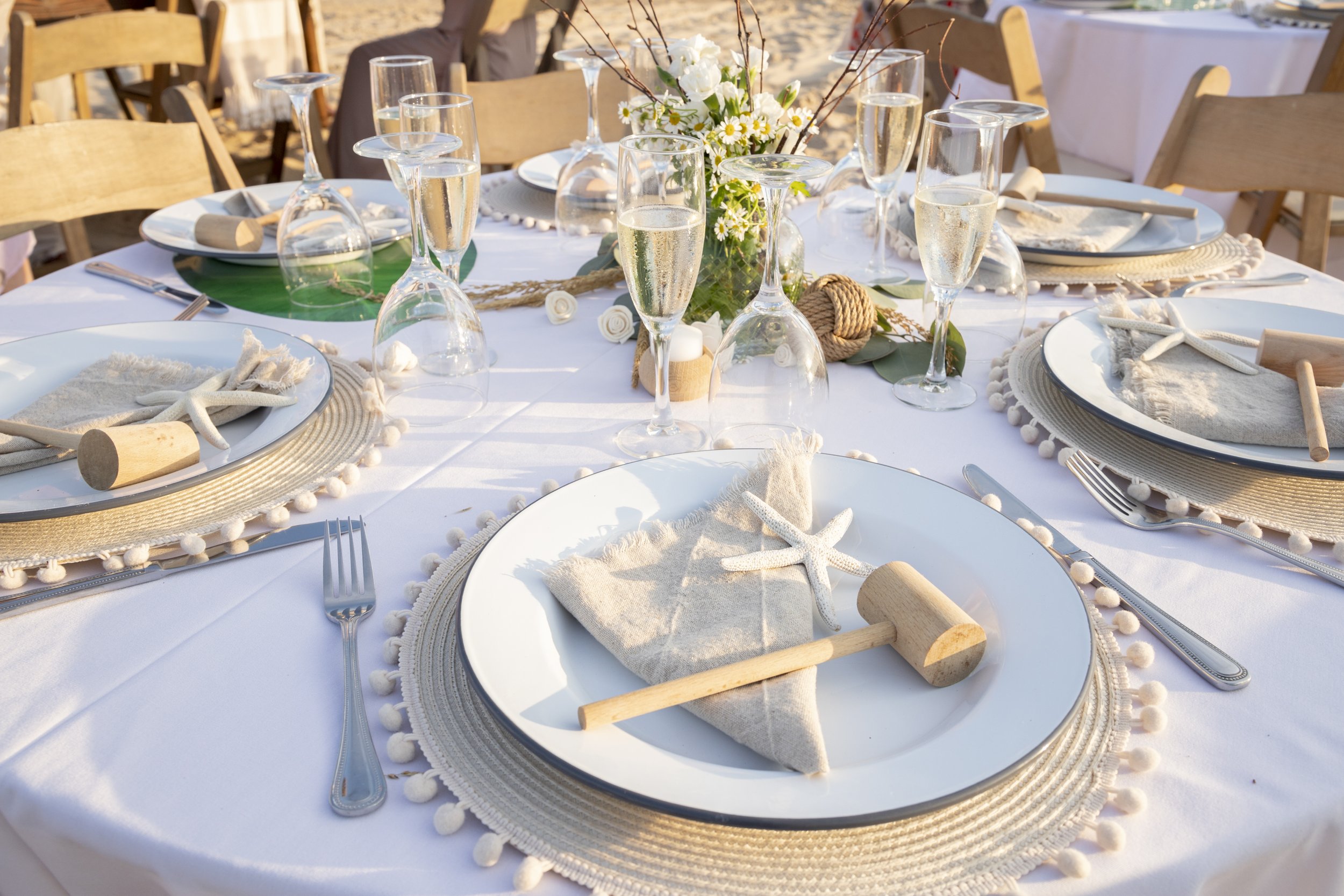 A beachside table setting with white tablecloths, starfish, seashells, and wooden mallets on each plate, with champagne glasses and a floral centerpiece.