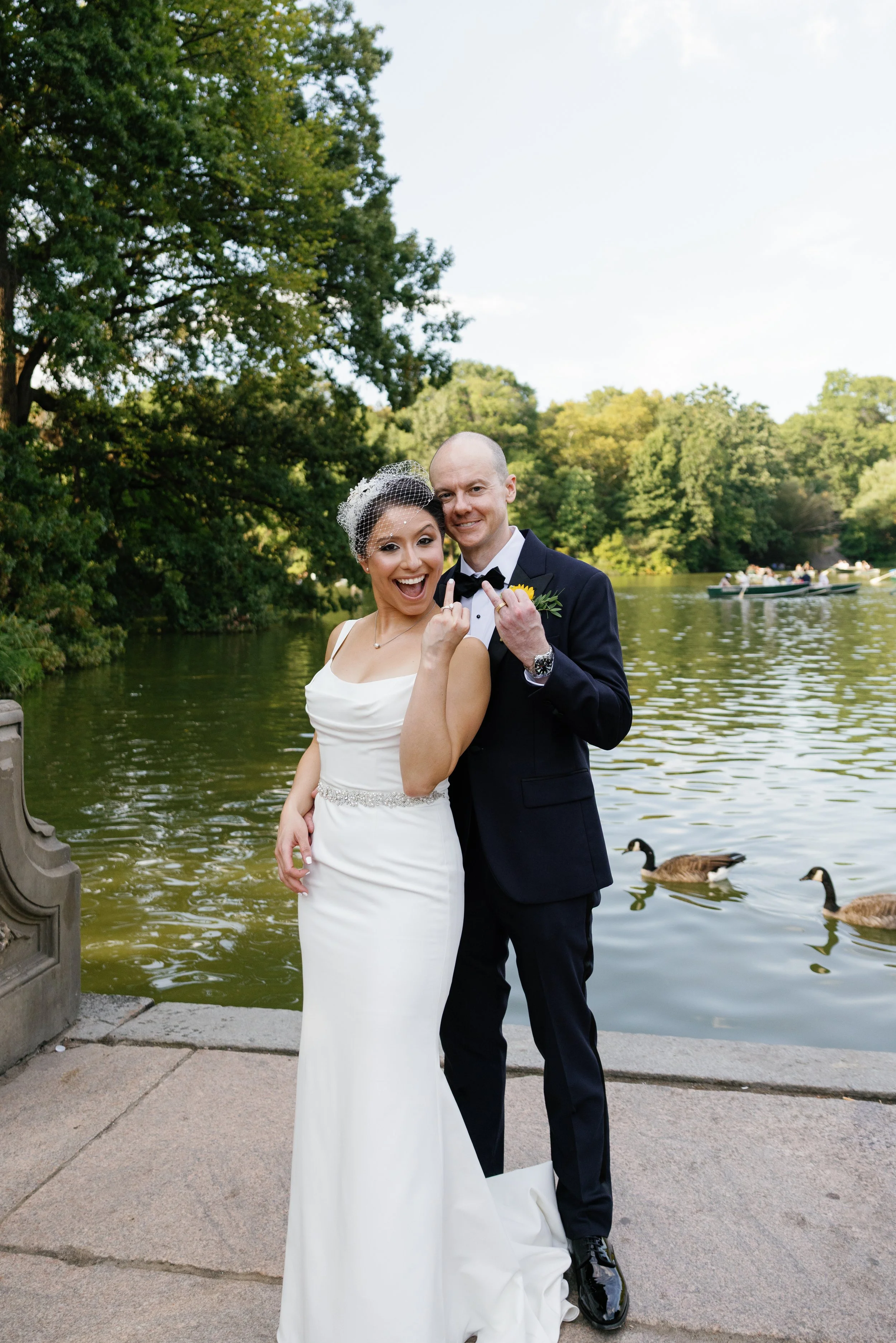 A newly married couple, a woman in a white wedding gown with a birdcage veil and a man in a black tuxedo with a yellow boutonniere, standing on a pathway by a river with ducks, surrounded by greenery, smiling and showing their wedding rings.